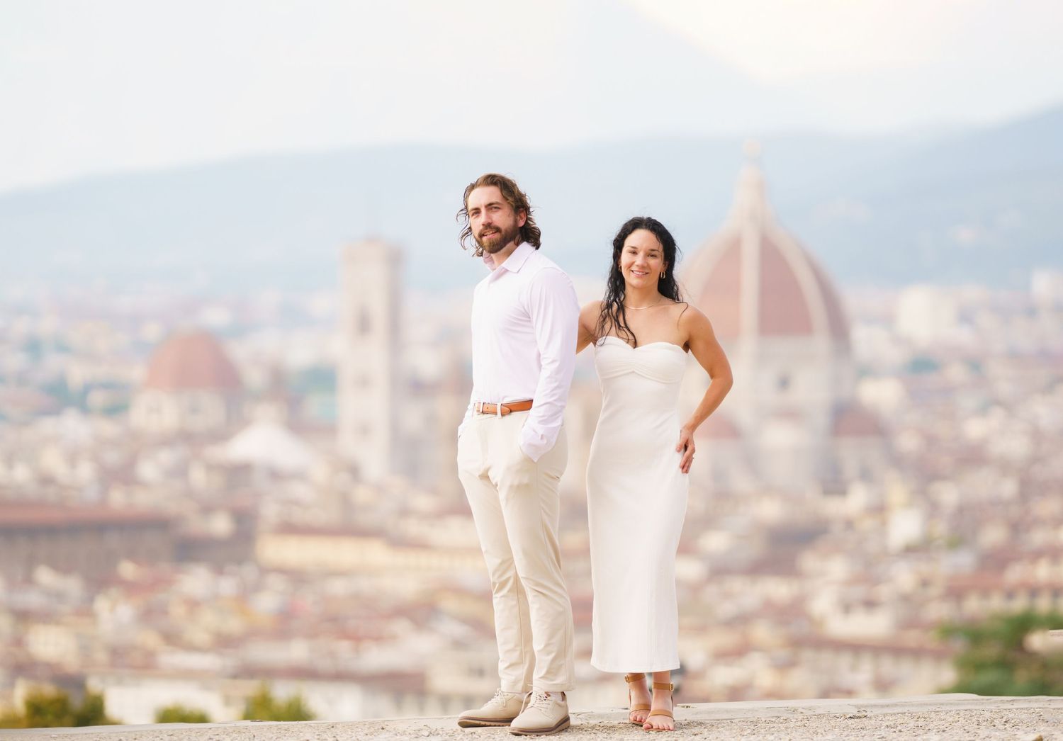Couple in white outfits holding hands overlooking a scenic European city with historic architecture in the background.