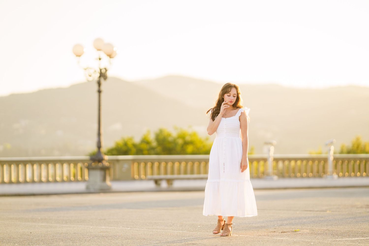 Woman in flowing white sundress stands on stone terrace at sunset with ornate lamp post and mountains in background.