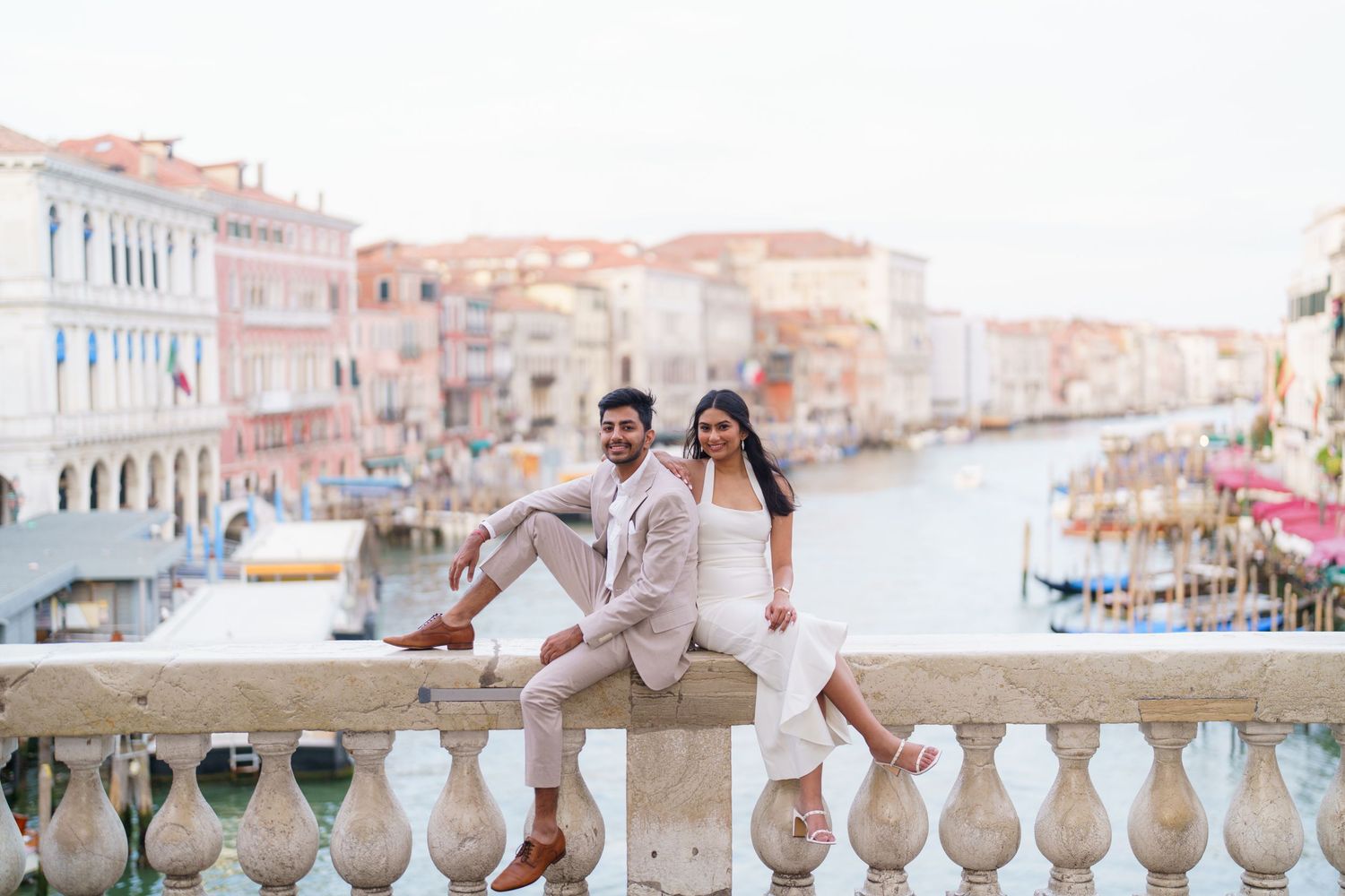 A romantic couple stands on a balcony overlooking the Venice canals and historic buildings during sunset.