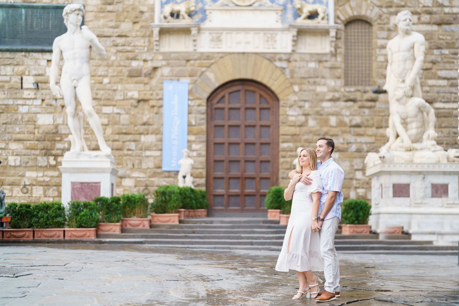 A couple in elegant white outfits poses together in front of a historic stone building with classical statues.