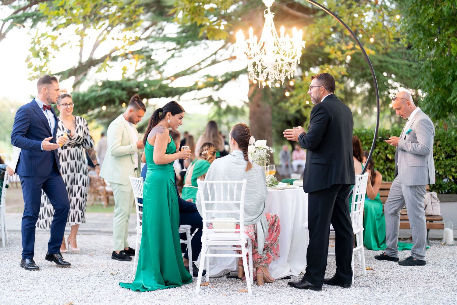 Wedding guests mingle and socialize during an outdoor reception beneath trees at sunset.