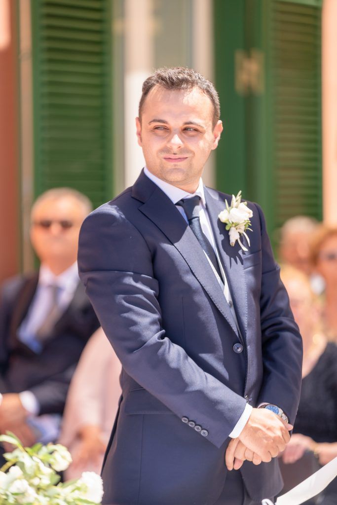 Wedding party members in navy suits gather on a terrace with green shutters.
