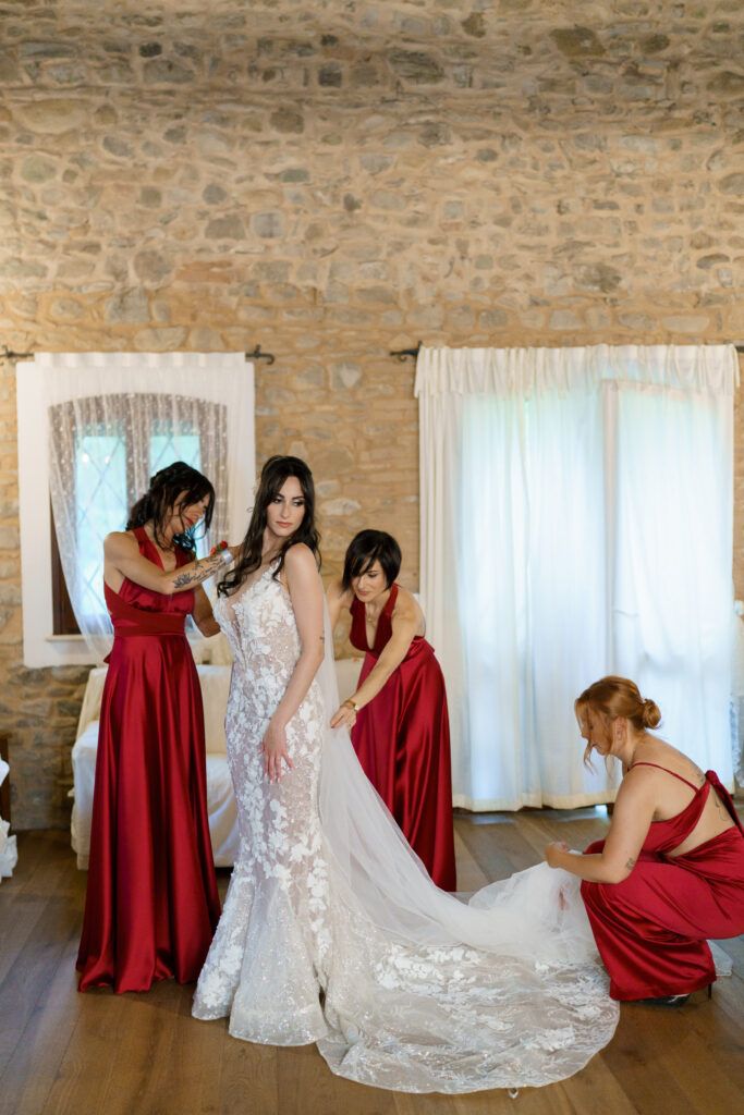 Wedding party in red dresses helping the bride with her dress in a stone-walled venue.