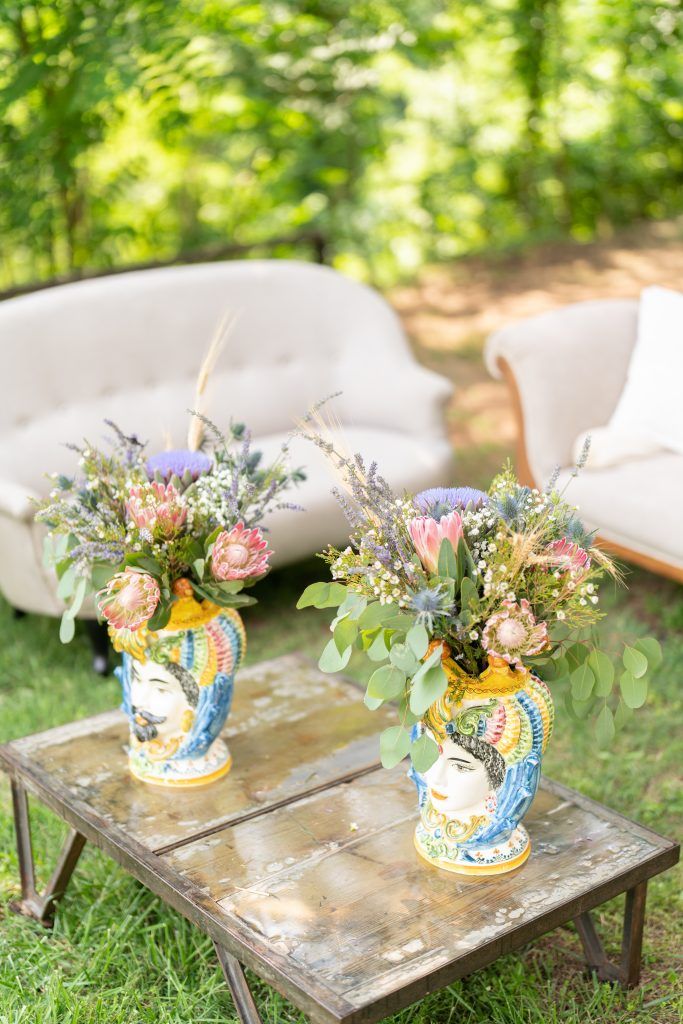 Rustic floral centerpieces in blue and yellow vases displayed on weathered wooden tables in an outdoor garden setting.
