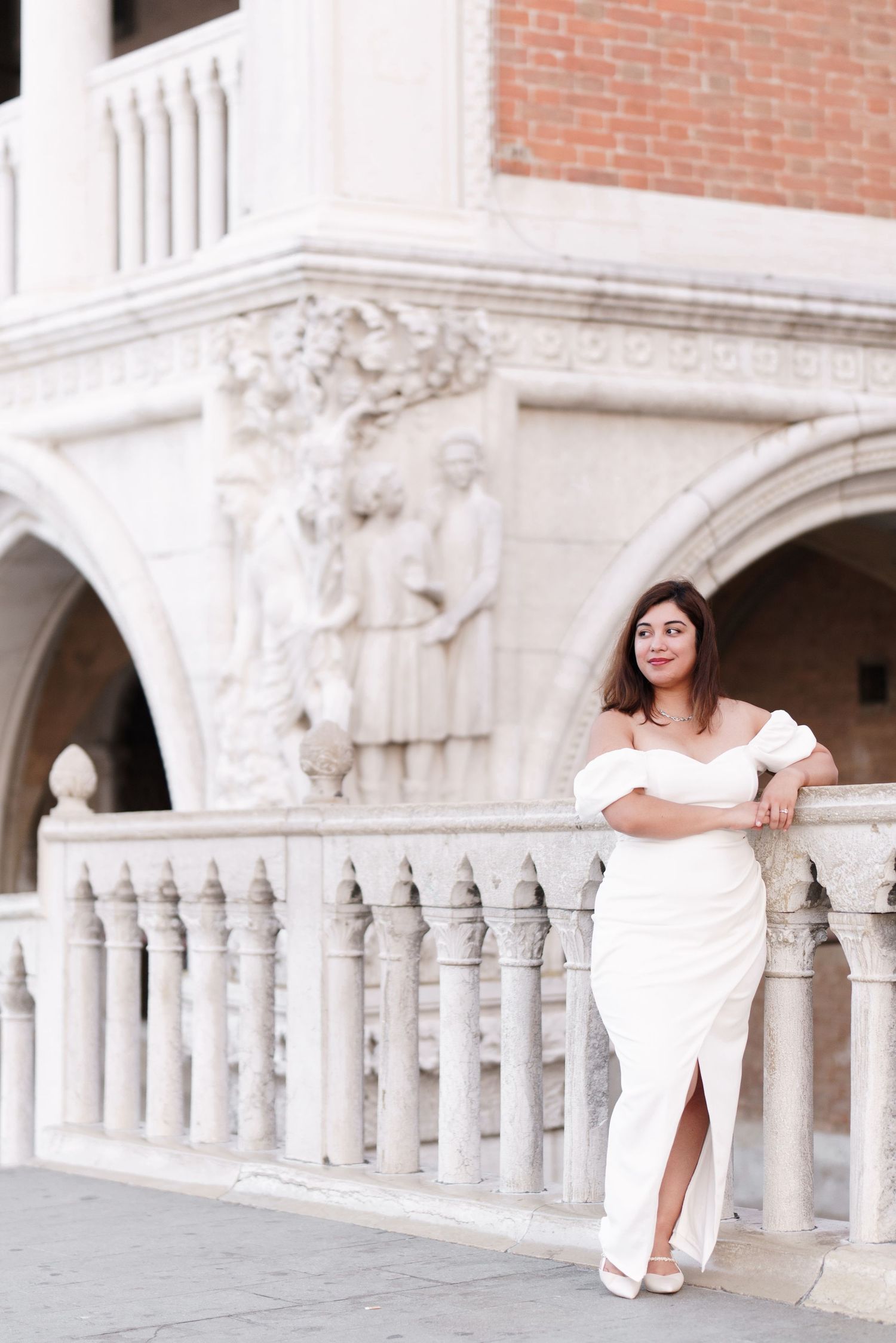 Woman in white dress poses elegantly against ornate Venetian architecture with classical columns and arches.