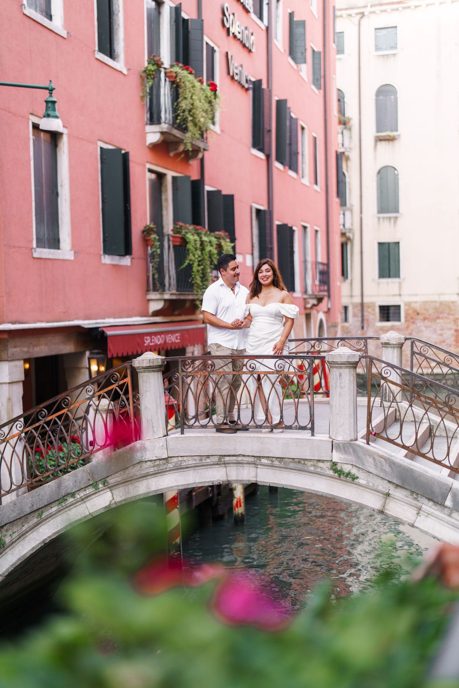 photographer venice A romantic scene on a small bridge over a Venetian canal with pink buildings and flowers in the foreground.