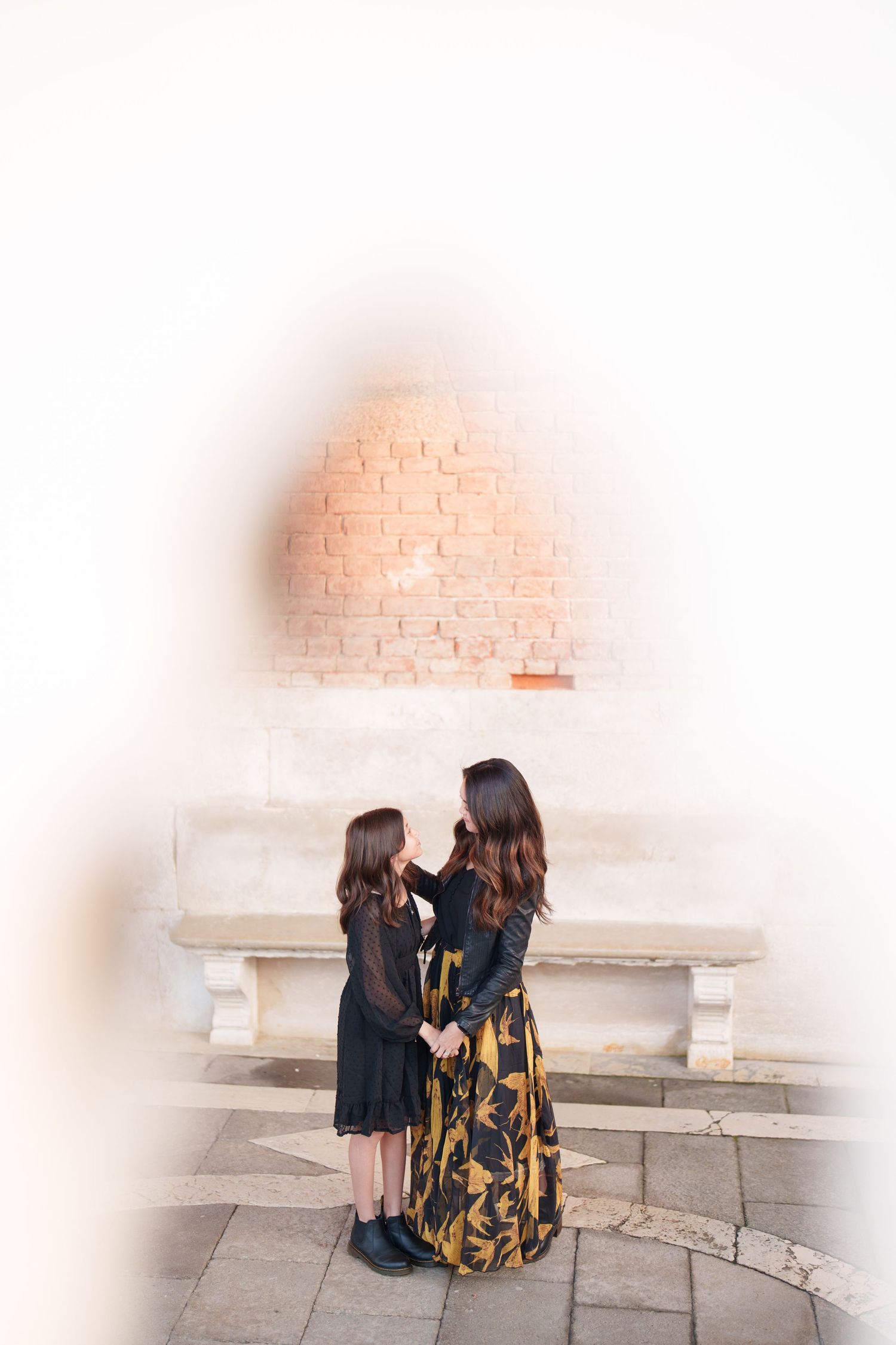 Two figures in dark dresses stand together on stone pavement against a light brick wall background.