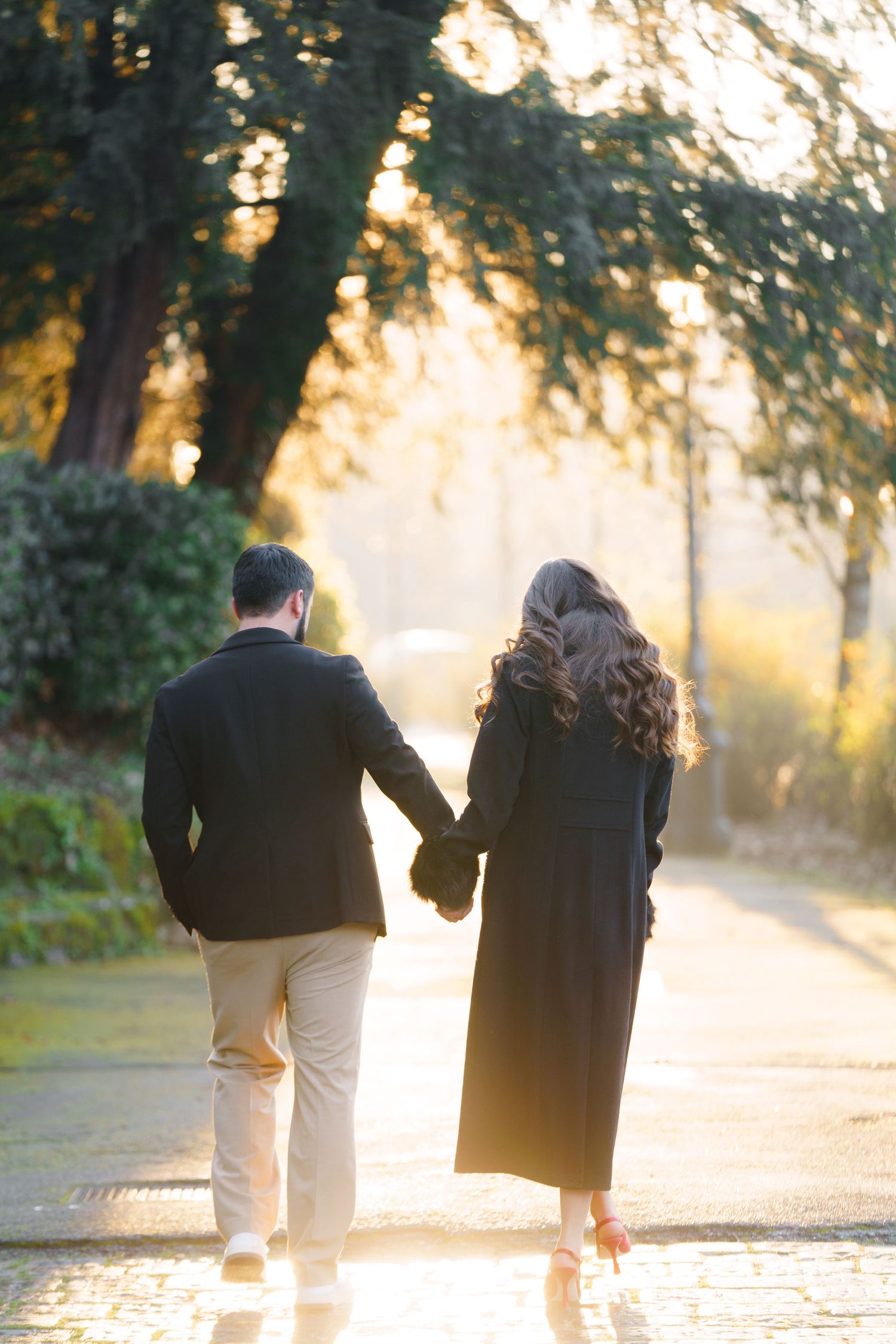 A couple walks hand in hand down a sunlit path through trees during golden hour.