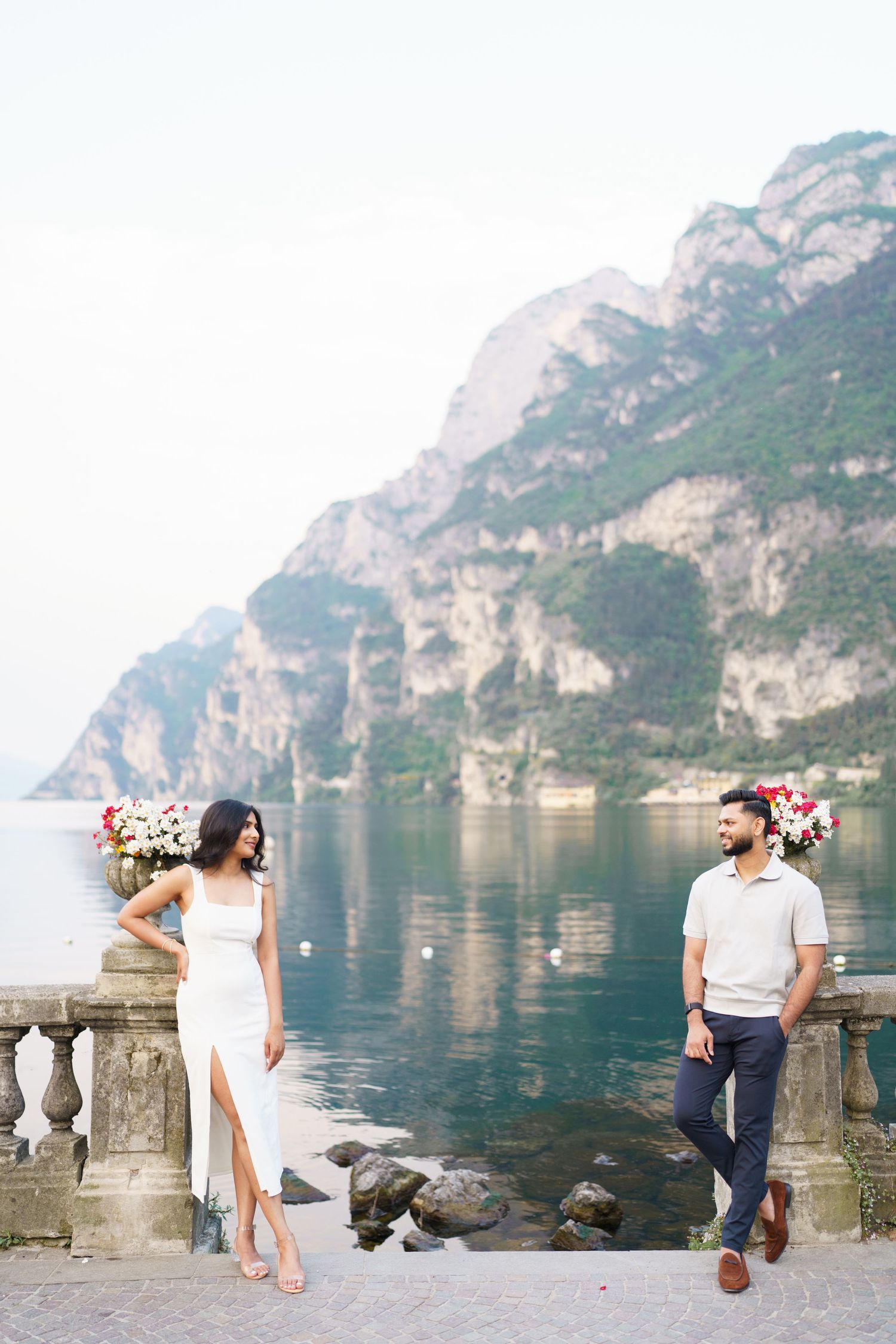 Two people in romantic attire pose on a stone balcony overlooking a stunning mountain lake in Italy.