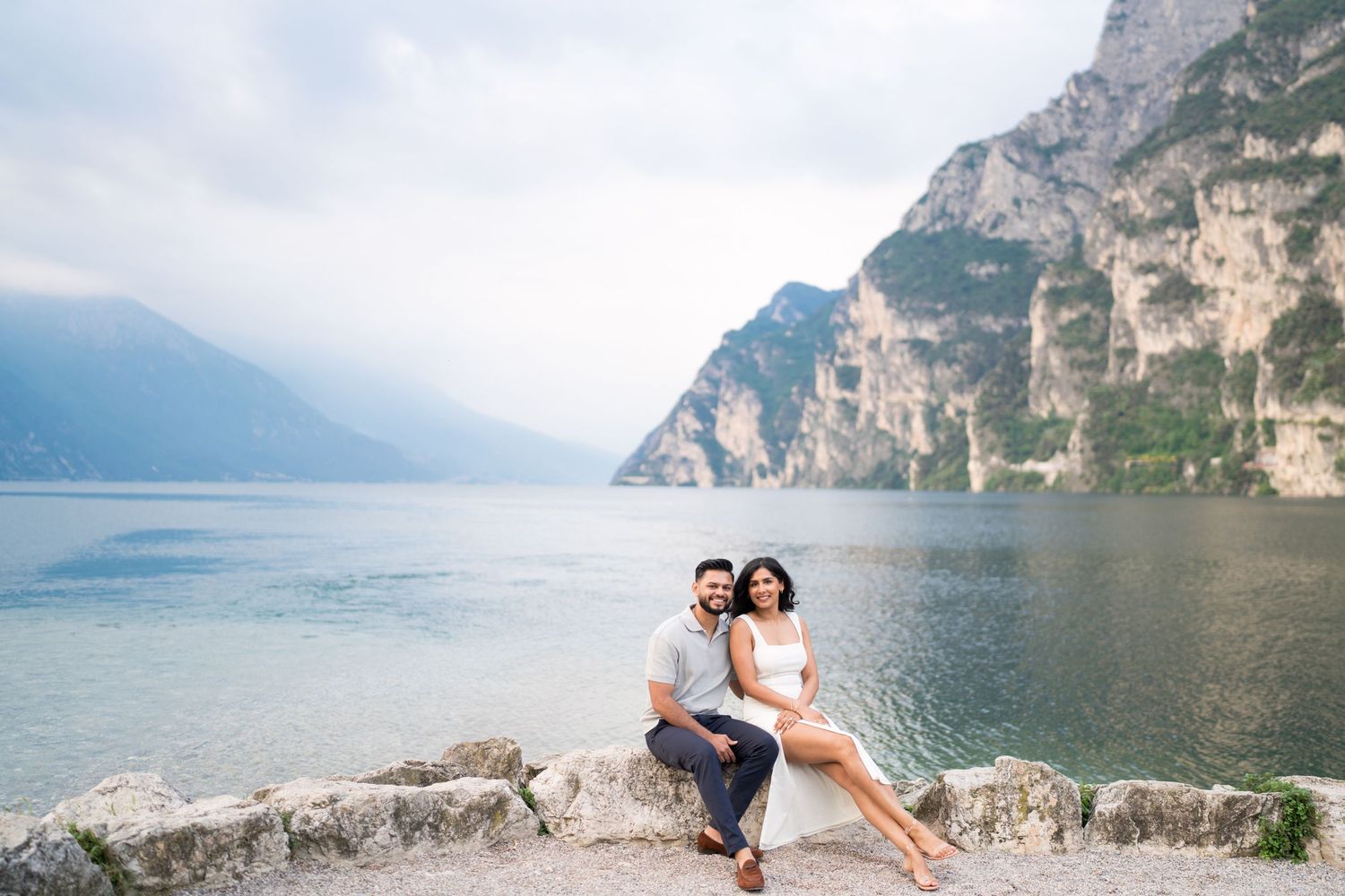 A couple sits together on rocky shore of Lake Garda in Italy with dramatic mountain cliffs in background.