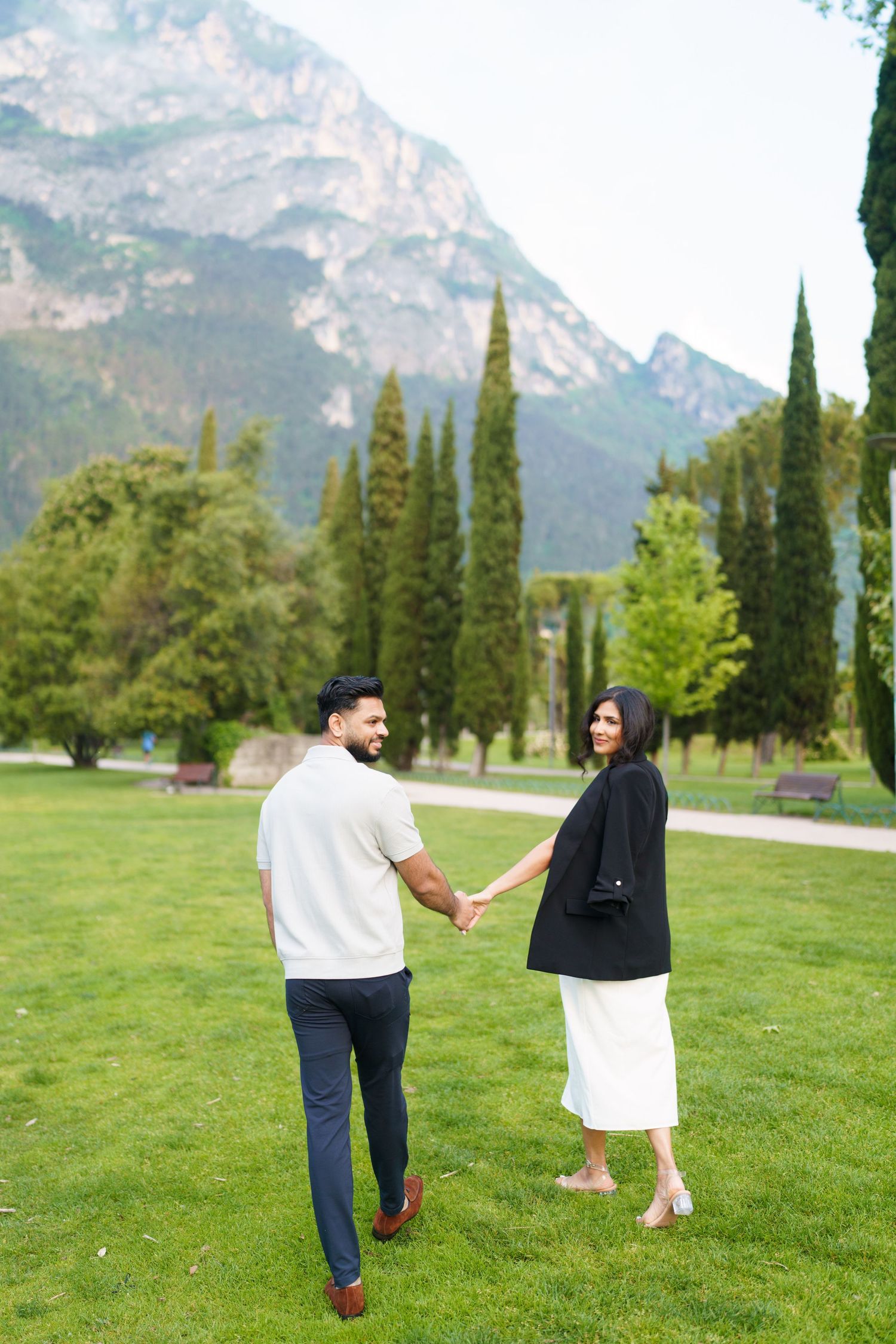 A couple walks together on a grassy field with tall cypress trees and majestic mountain peaks in the background.