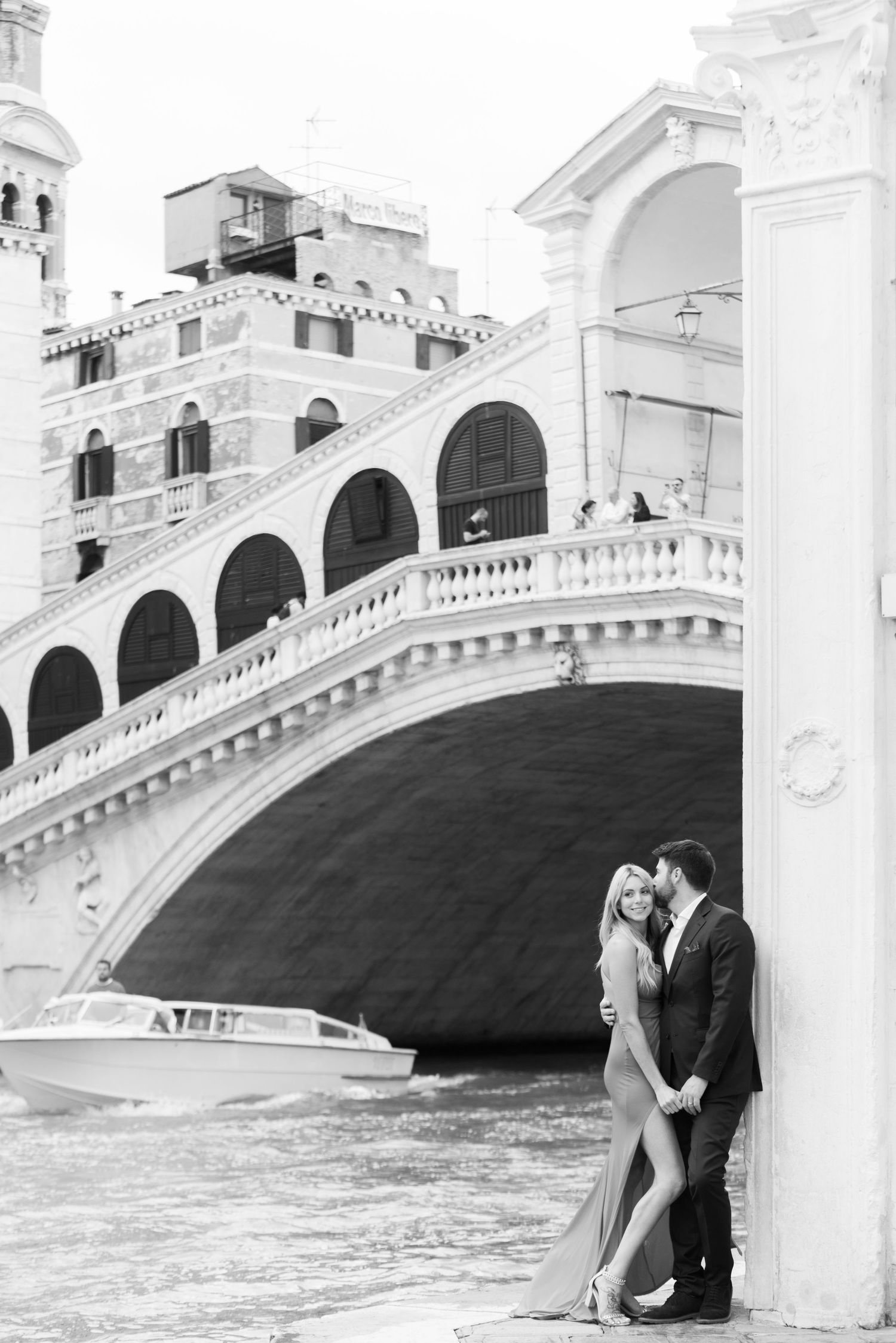 Romantic black and white photo near the iconic Rialto Bridge in Venice with a couple embracing by the canal.