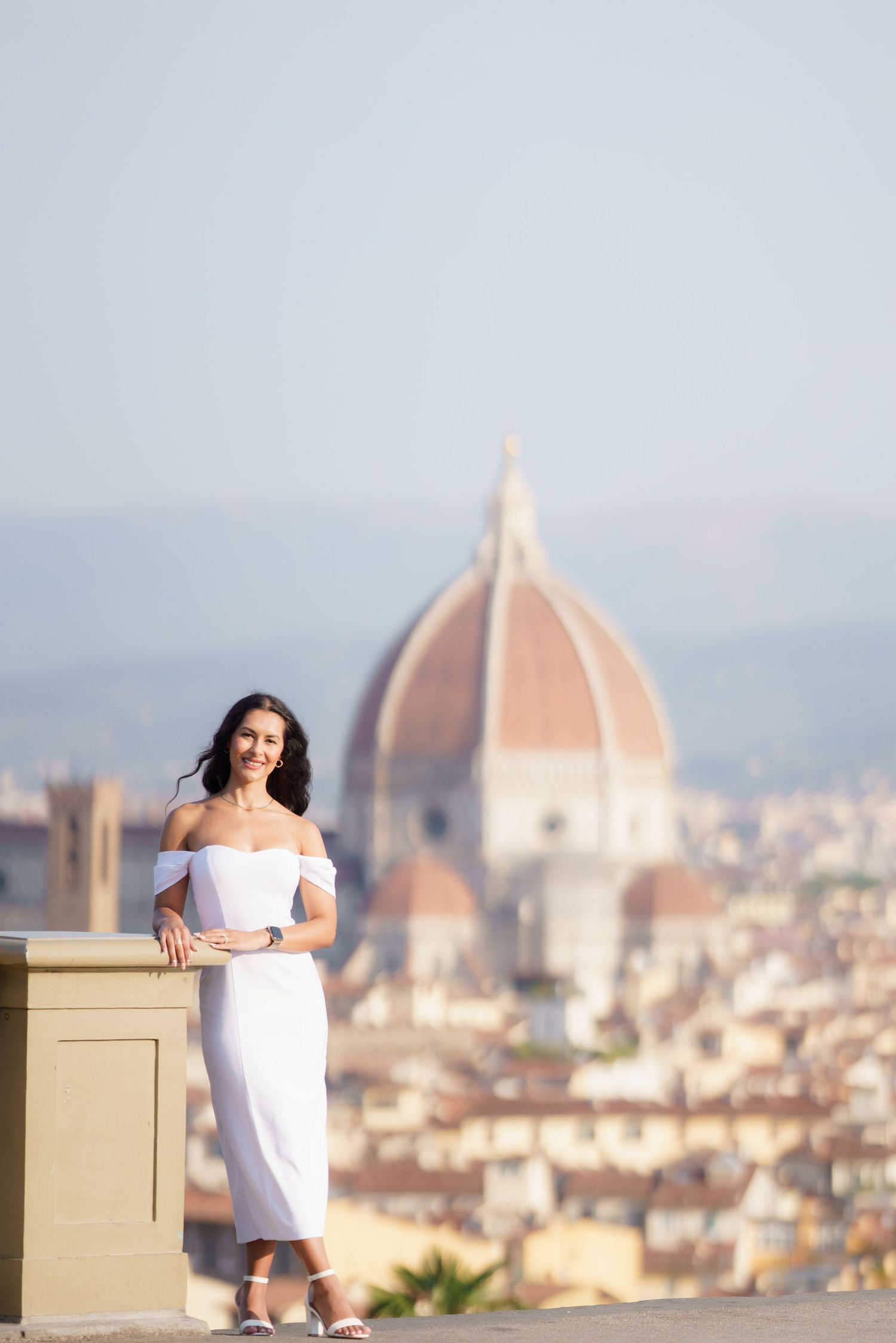 Woman in white dress poses at scenic viewpoint overlooking Florence with iconic Duomo cathedral dome in background.