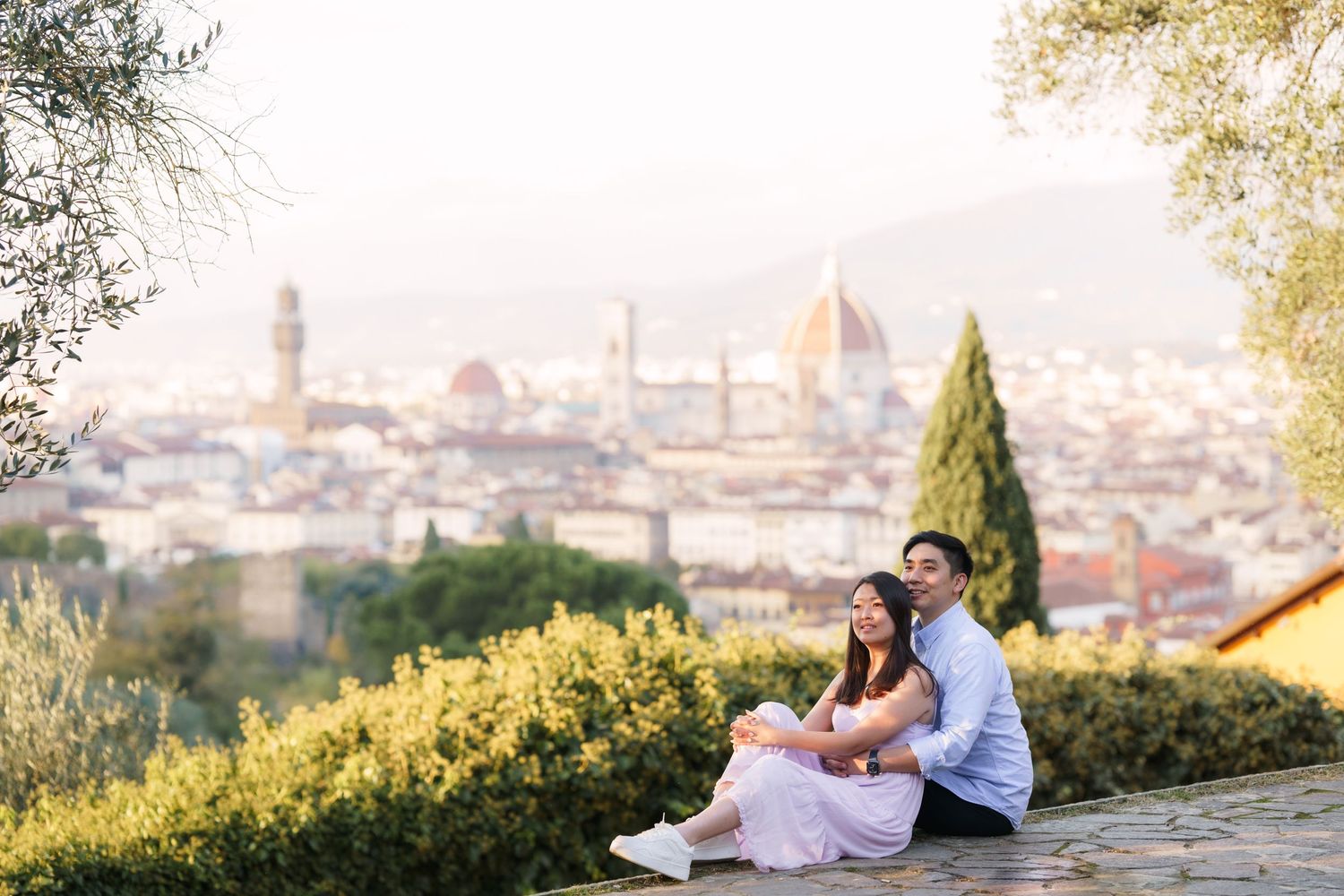 A couple sits on a hillside overlooking Florence, Italy with the Duomo cathedral visible in the cityscape below.