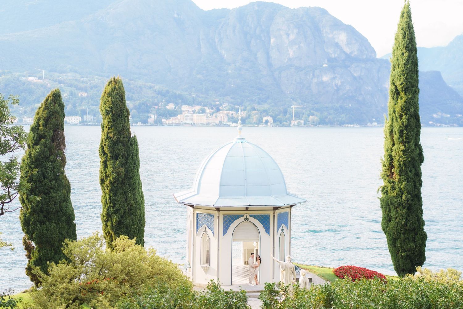 A white domed gazebo overlooking Lake Como with tall cypress trees and mountains in the background.
