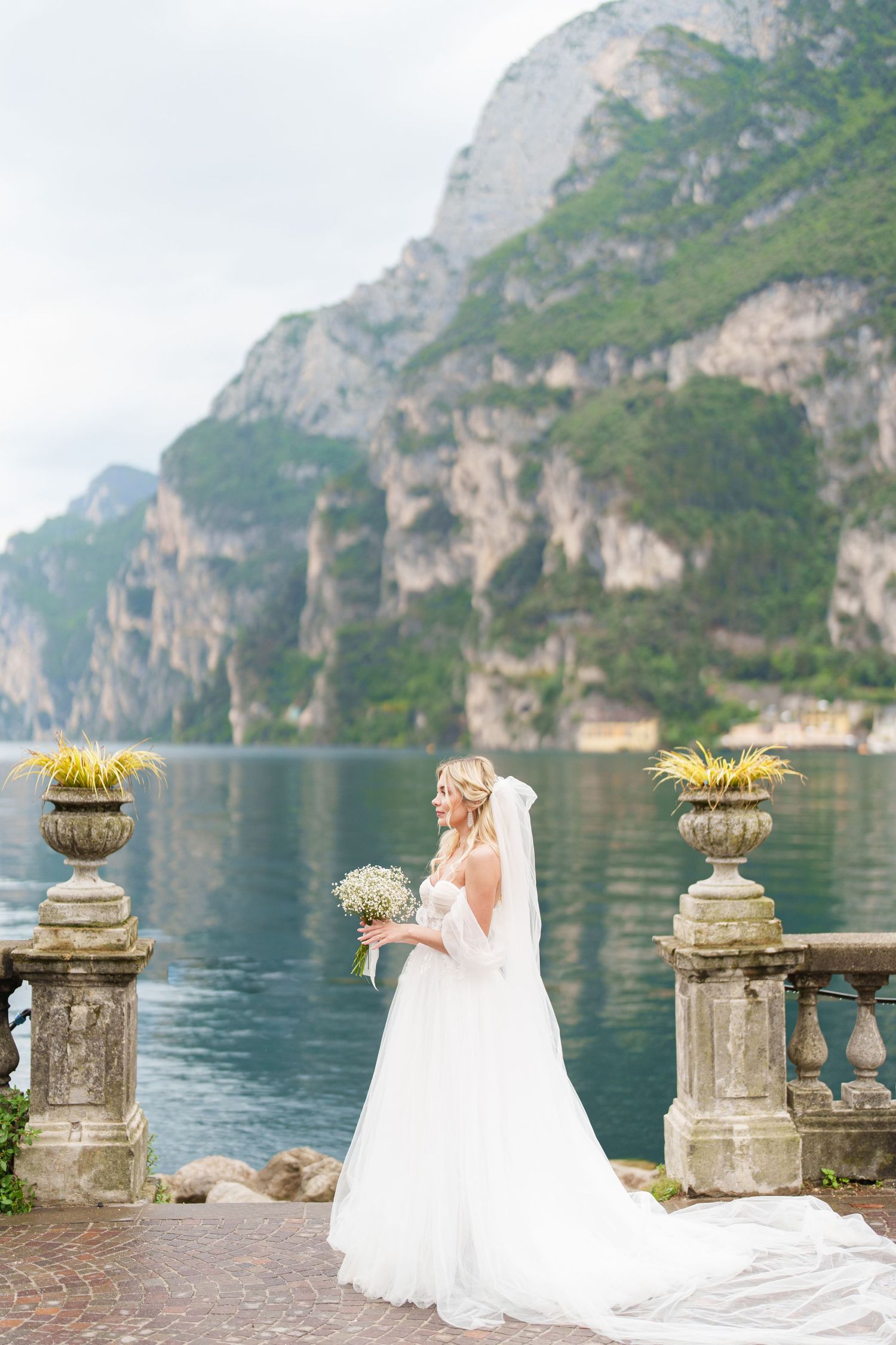 Bride in flowing white gown poses along scenic Lake Garda with mountain cliffs and stone balustrade.