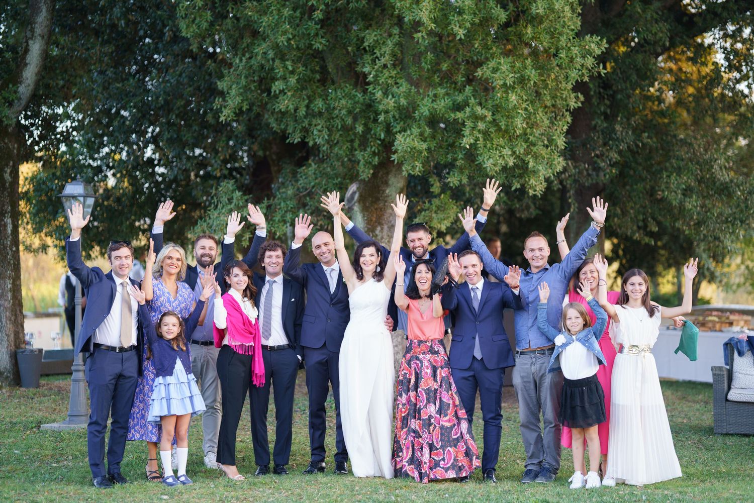 Wedding guests and party members pose energetically with arms raised under trees.