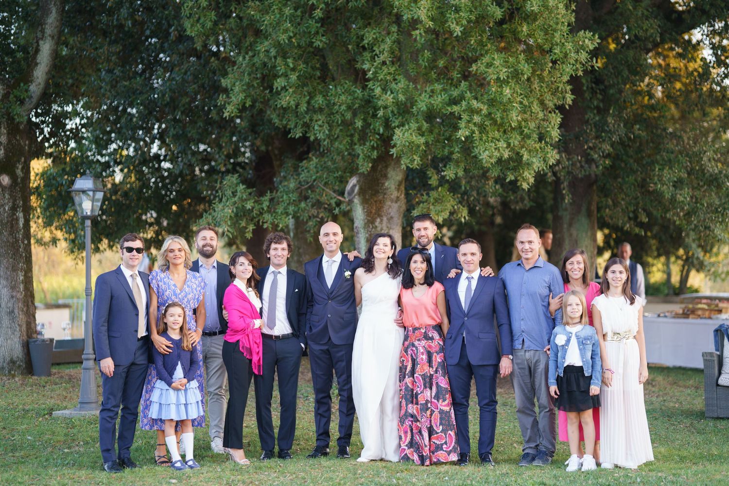 Wedding party group photo with guests standing together outdoors under large trees at sunset.