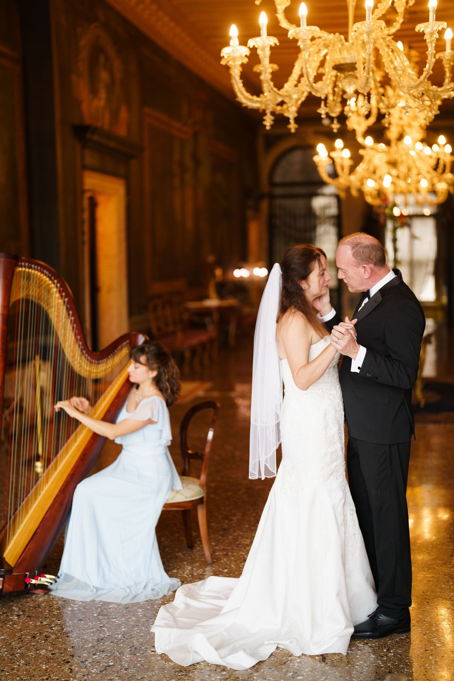 Elegant wedding scene in ornate ballroom with harpist playing under golden chandeliers as couple dances.