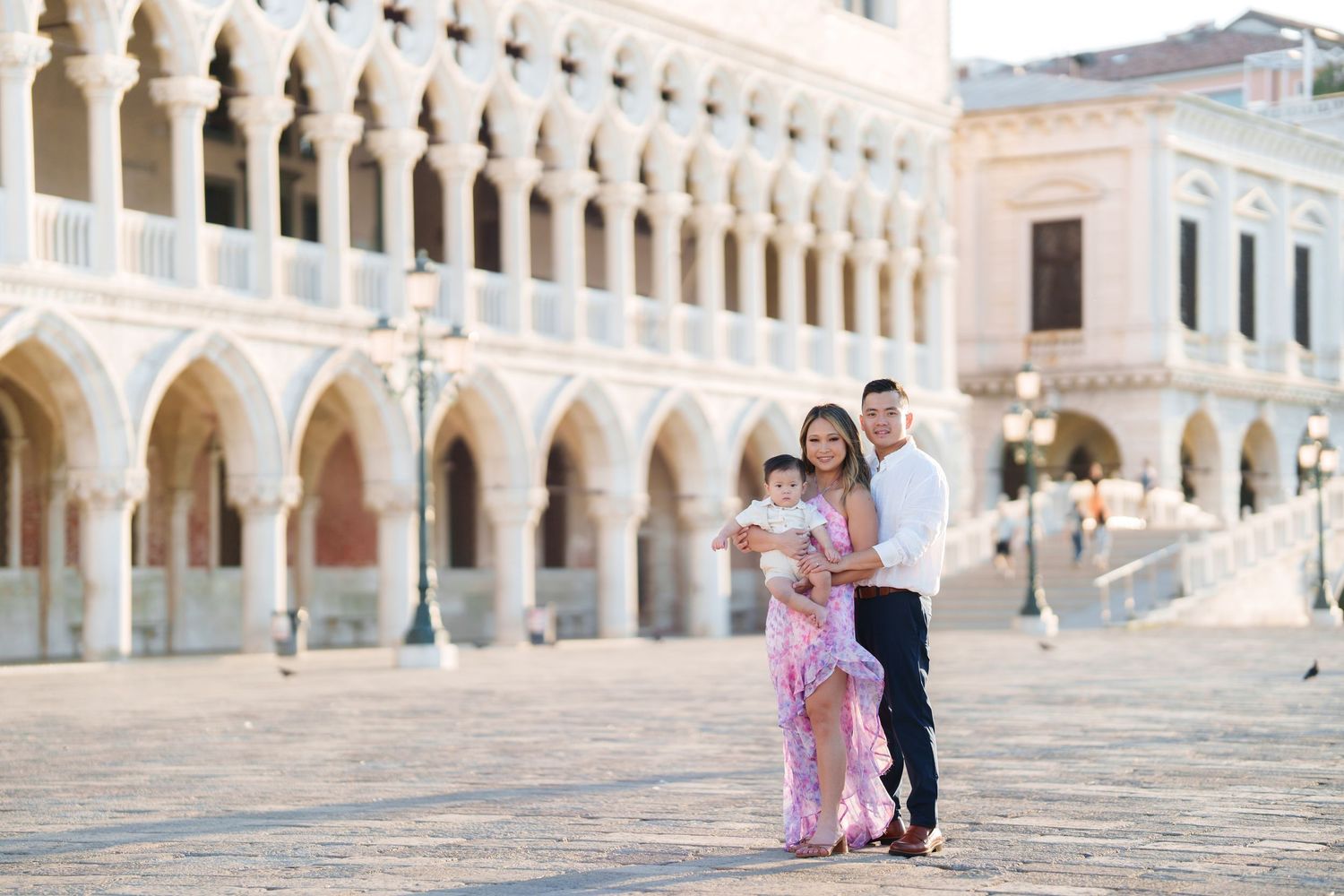 Family of three posing in a sunlit Venetian piazza, mother in pink floral gown holding baby, father in white shirt.