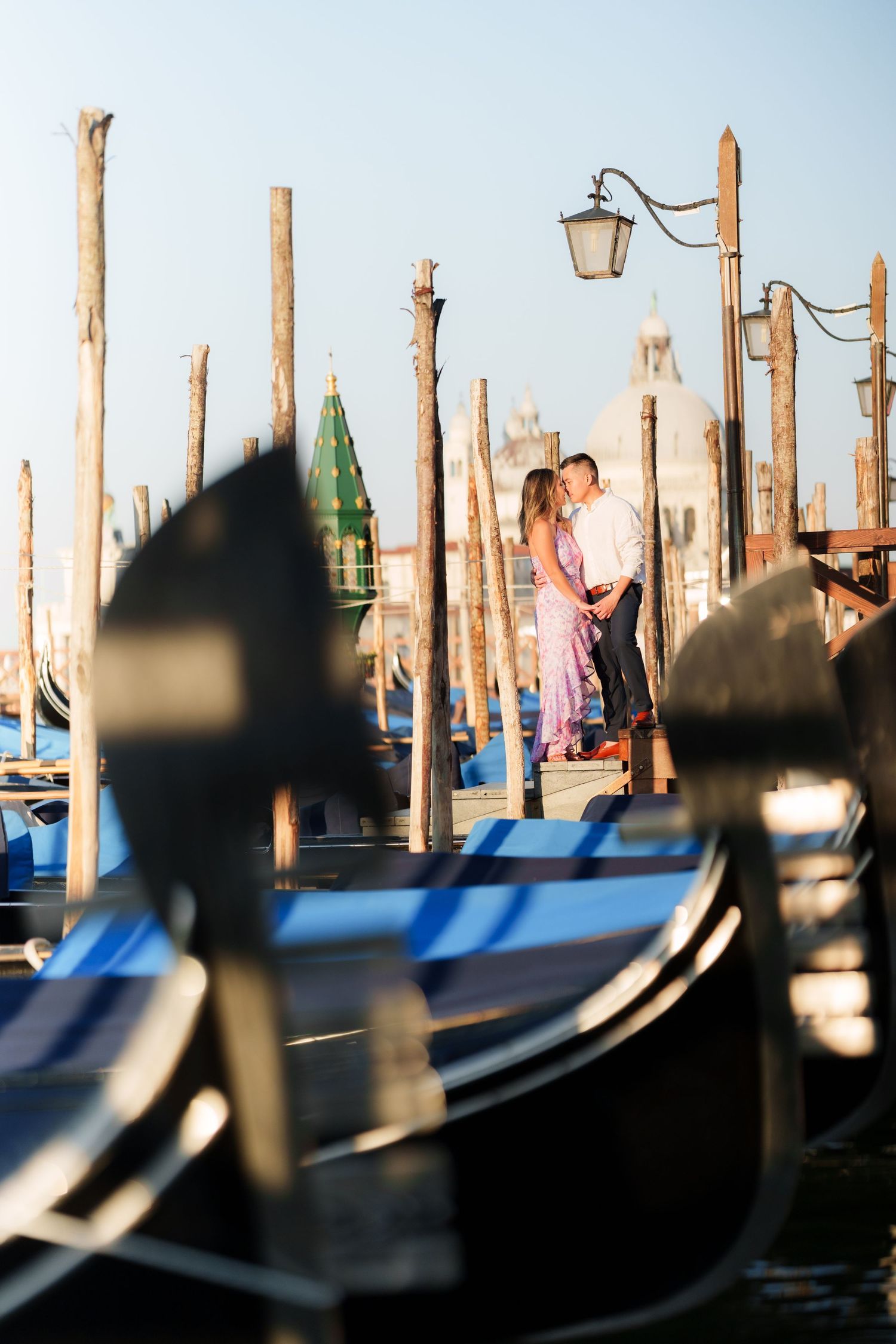 Romantic couple embracing among gondolas and wooden poles at Venice waterfront with church dome in background.