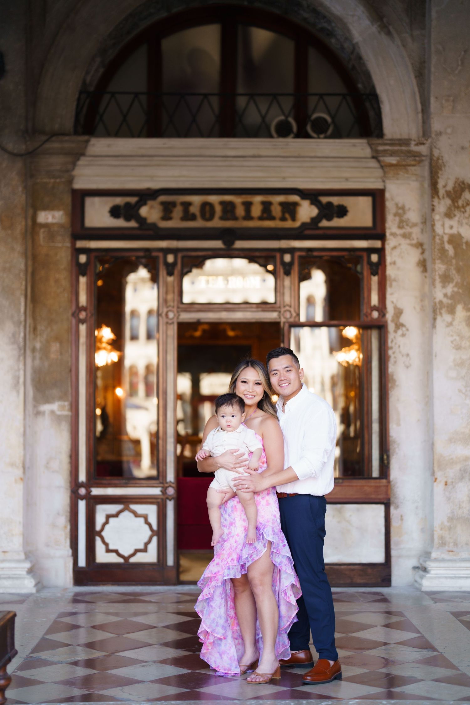A smiling family poses with their baby outside Café Florian in Venice, Italy, wearing elegant attire.