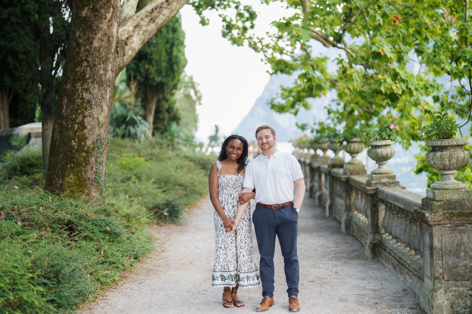 Smiling interracial couple standing together on a tree-lined garden path with stone balustrade.