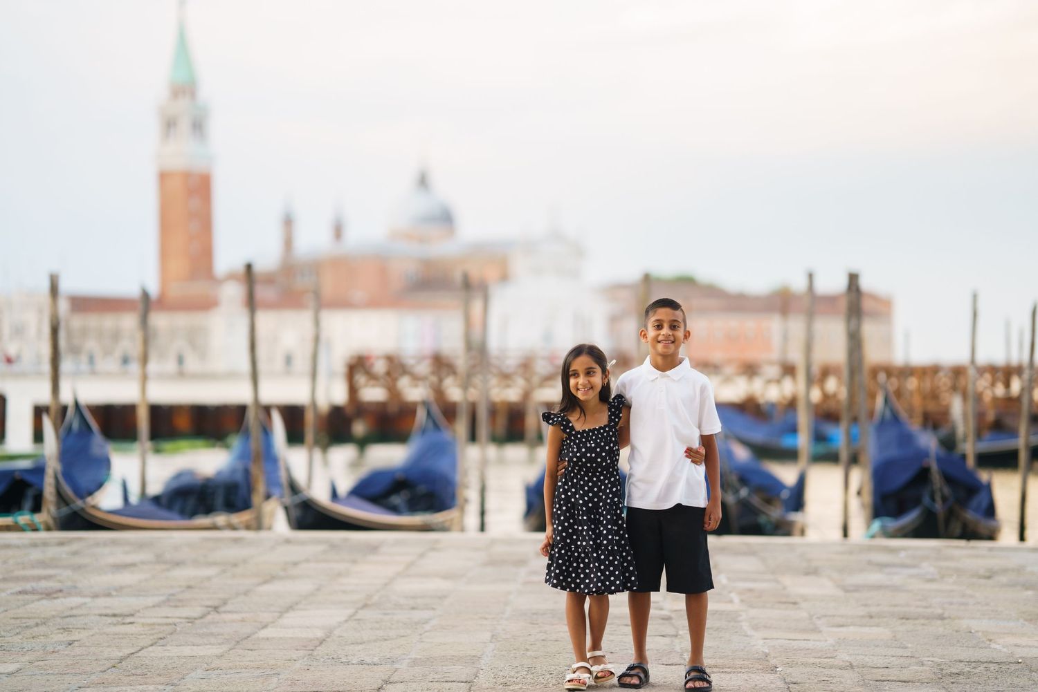 Two children posing together on a Venice waterfront with gondolas and San Giorgio Maggiore in the background.