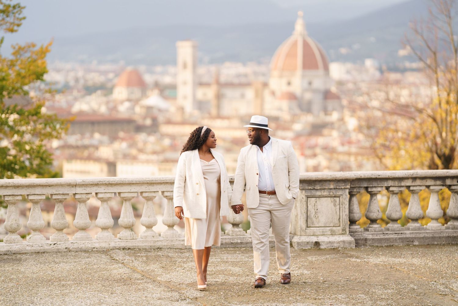 Couple in white attire holding hands at Piazzale Michelangelo with Florence cathedral skyline backdrop.