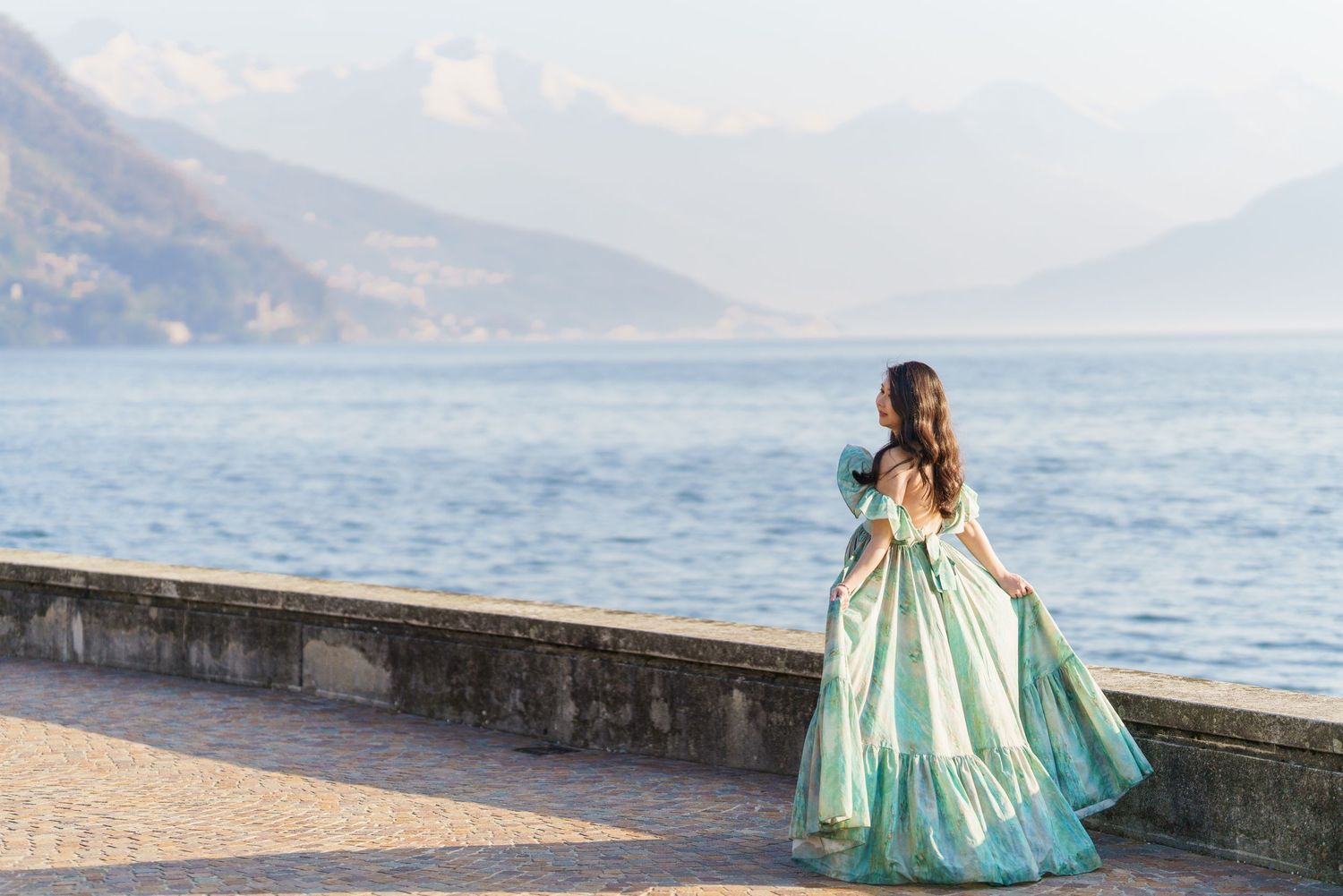 A woman in an elegant teal ball gown stands on a lakeside promenade with misty mountains in the background.