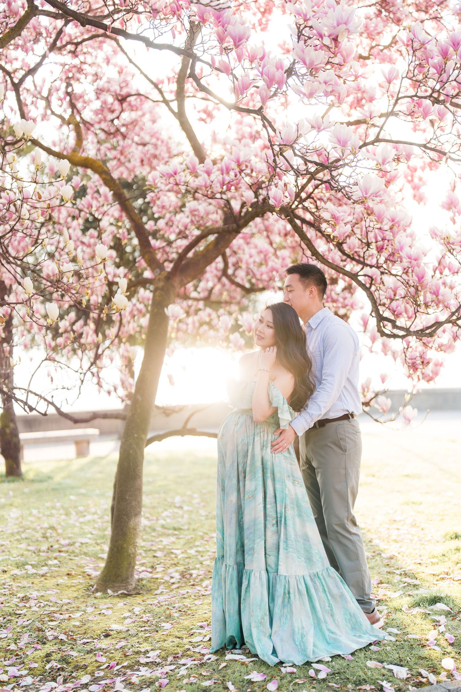 Couple embracing under pink magnolia tree in bloom, woman in teal floral gown, soft golden backlight, spring garden setting.