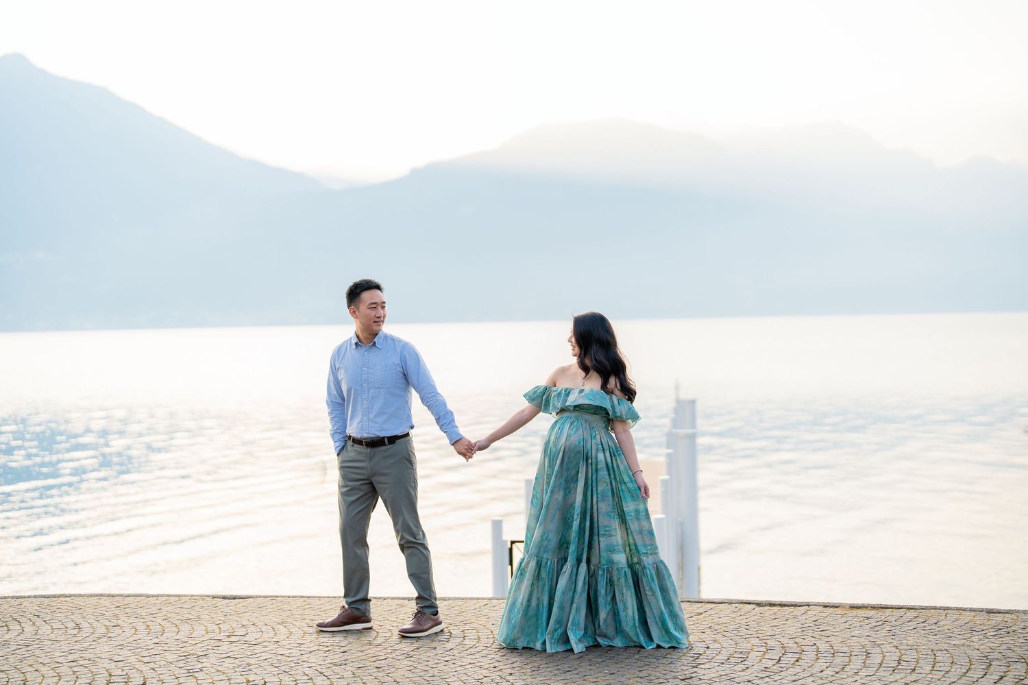 Couple holding hands by a lakeside with mountains, woman in teal gown, man in blue shirt, romantic outdoor photo.