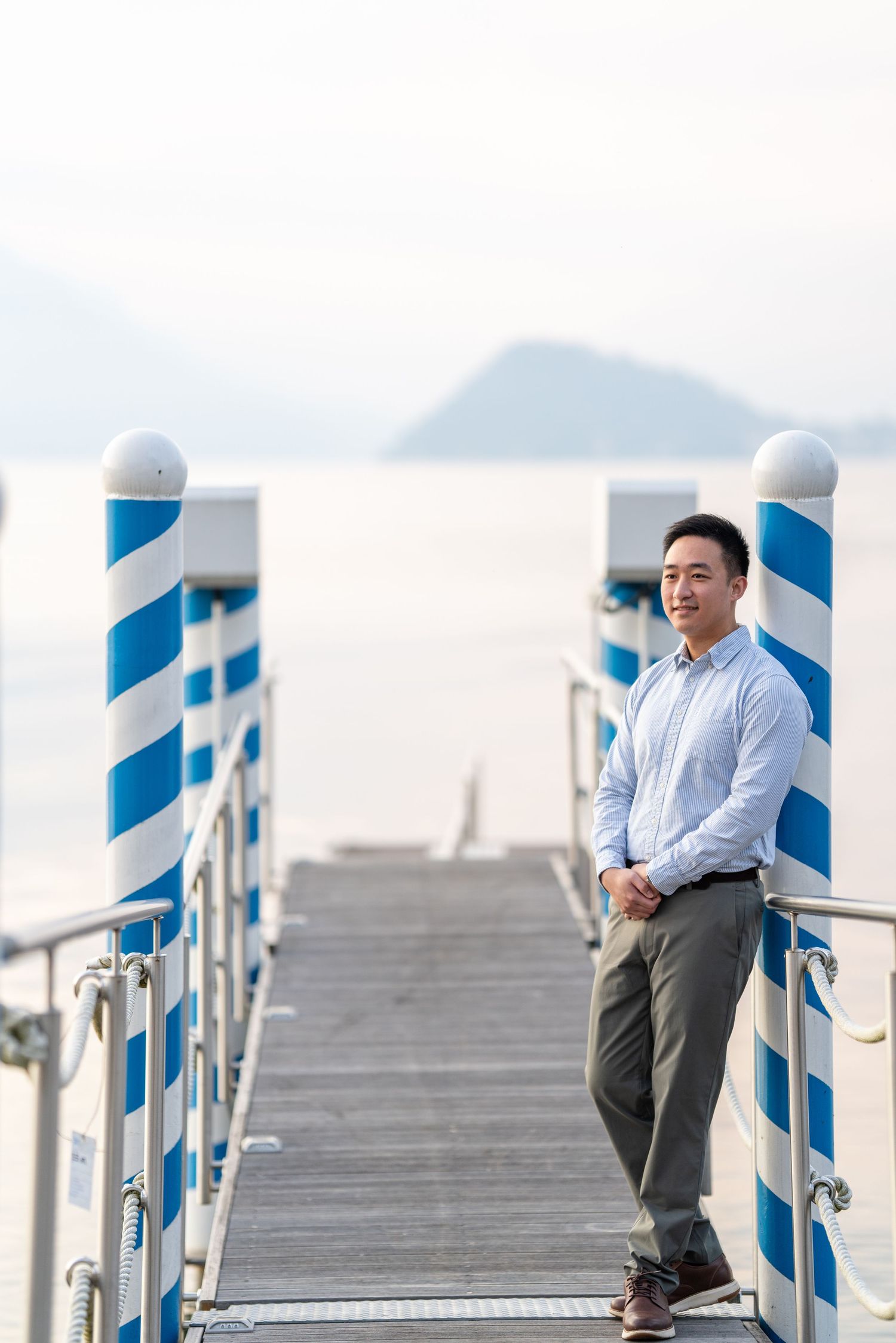 Young Asian man in blue shirt poses confidently on a waterfront pier with blue and white striped poles and misty mountains.