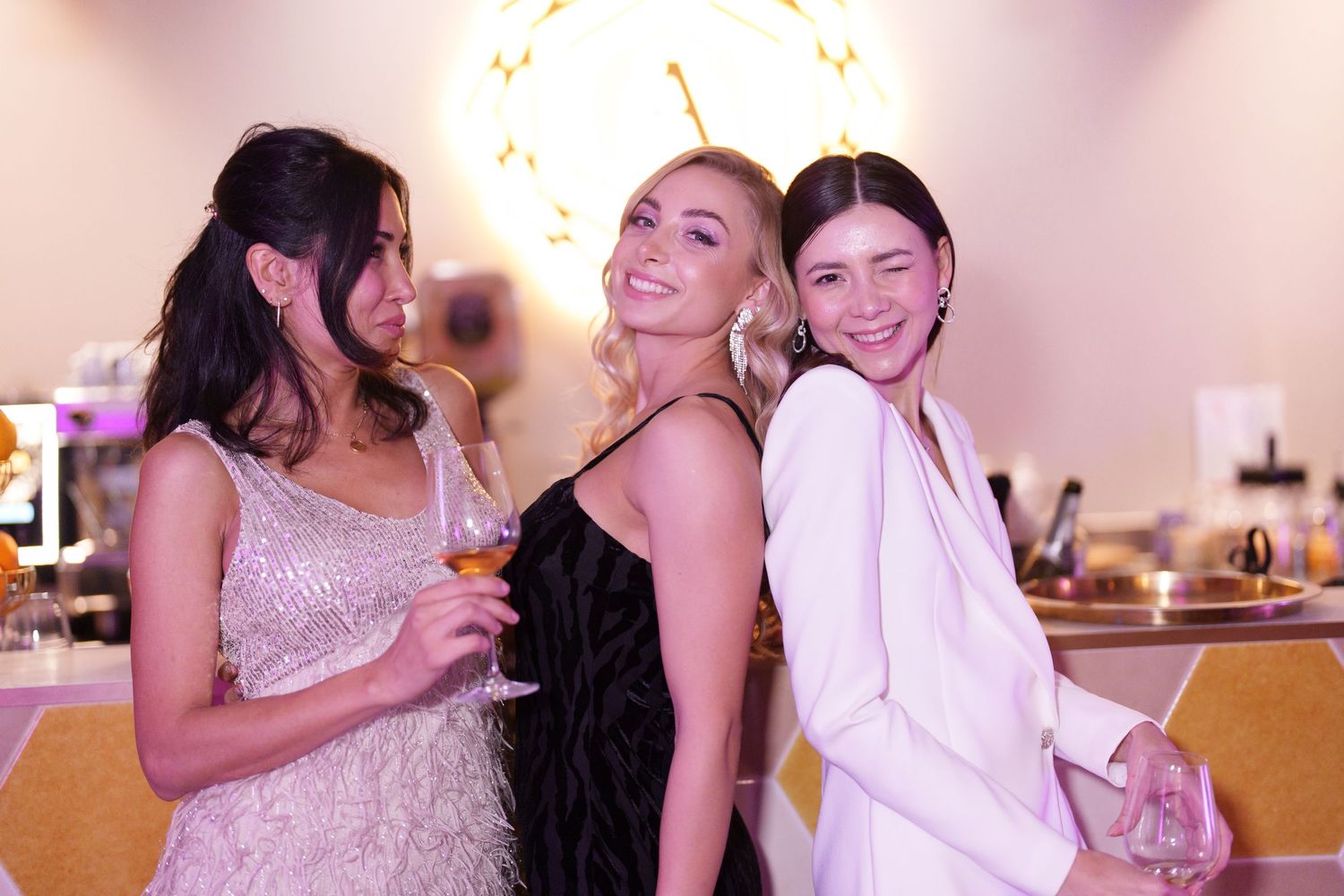 Three women in elegant dresses smiling and posing together at a glamorous party event.