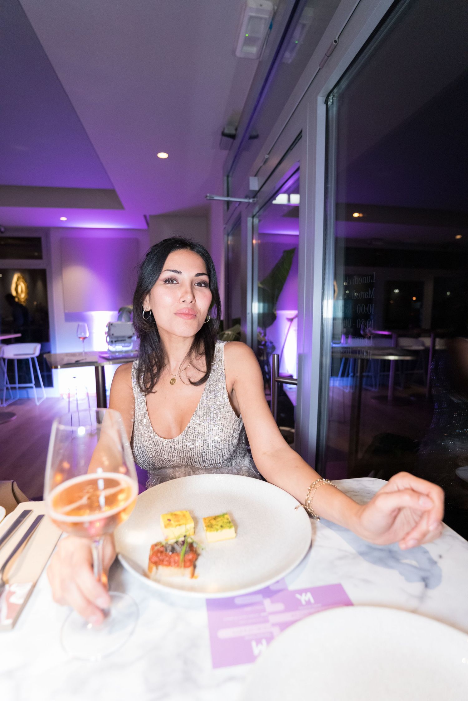 Woman in silver dress dining at upscale restaurant with wine and gourmet appetizers, purple ambient lighting.