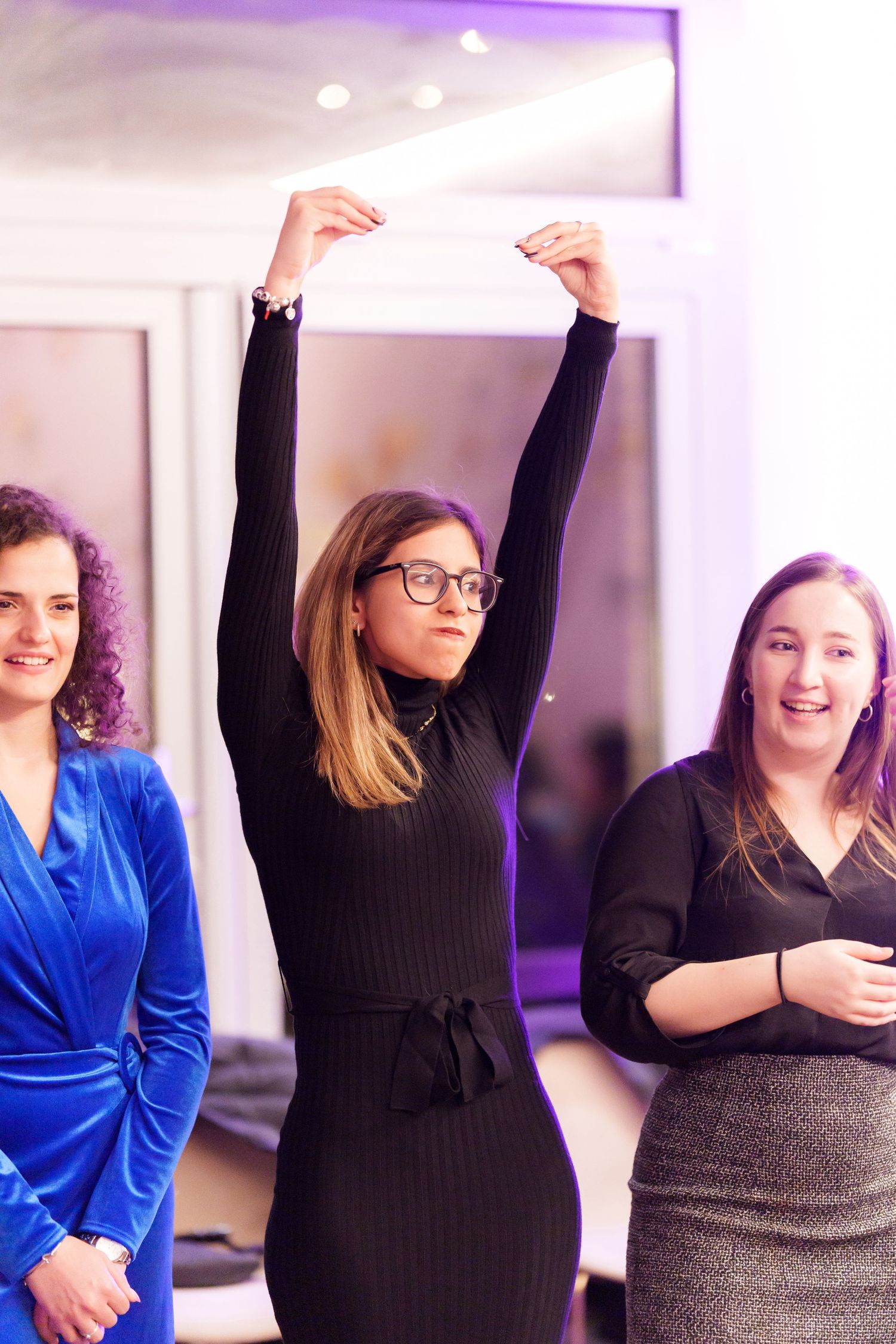 Three women at an indoor event, center woman in black dress raises arms triumphantly, flanked by smiling companions.