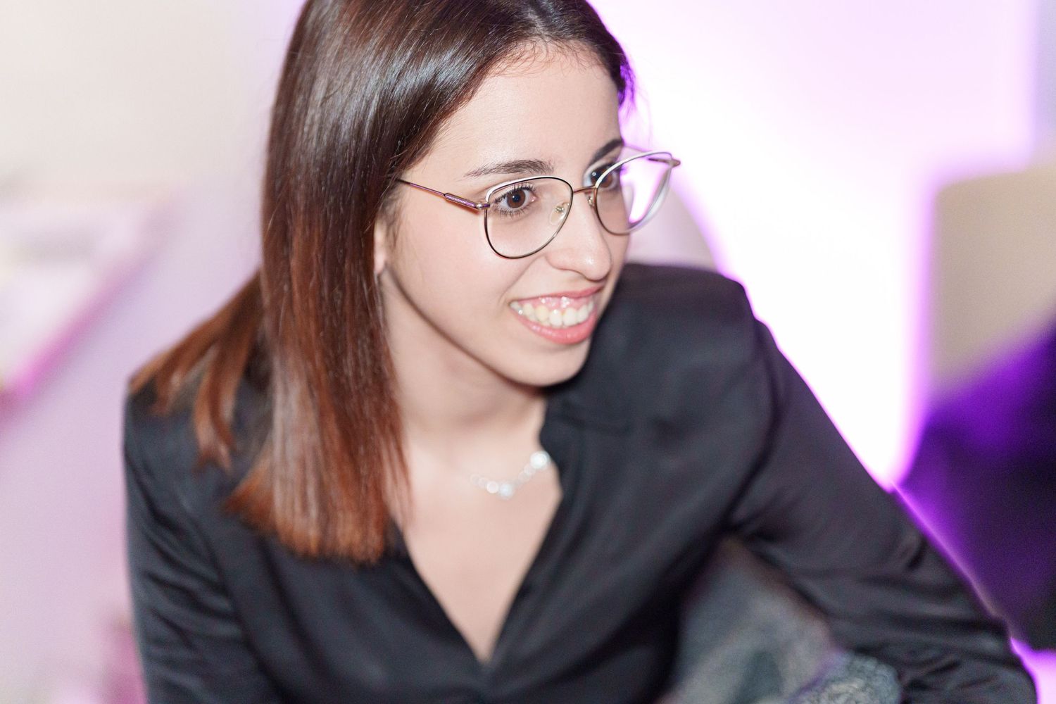 Young woman with glasses and dark hair smiling, wearing a black blazer in a softly lit environment.