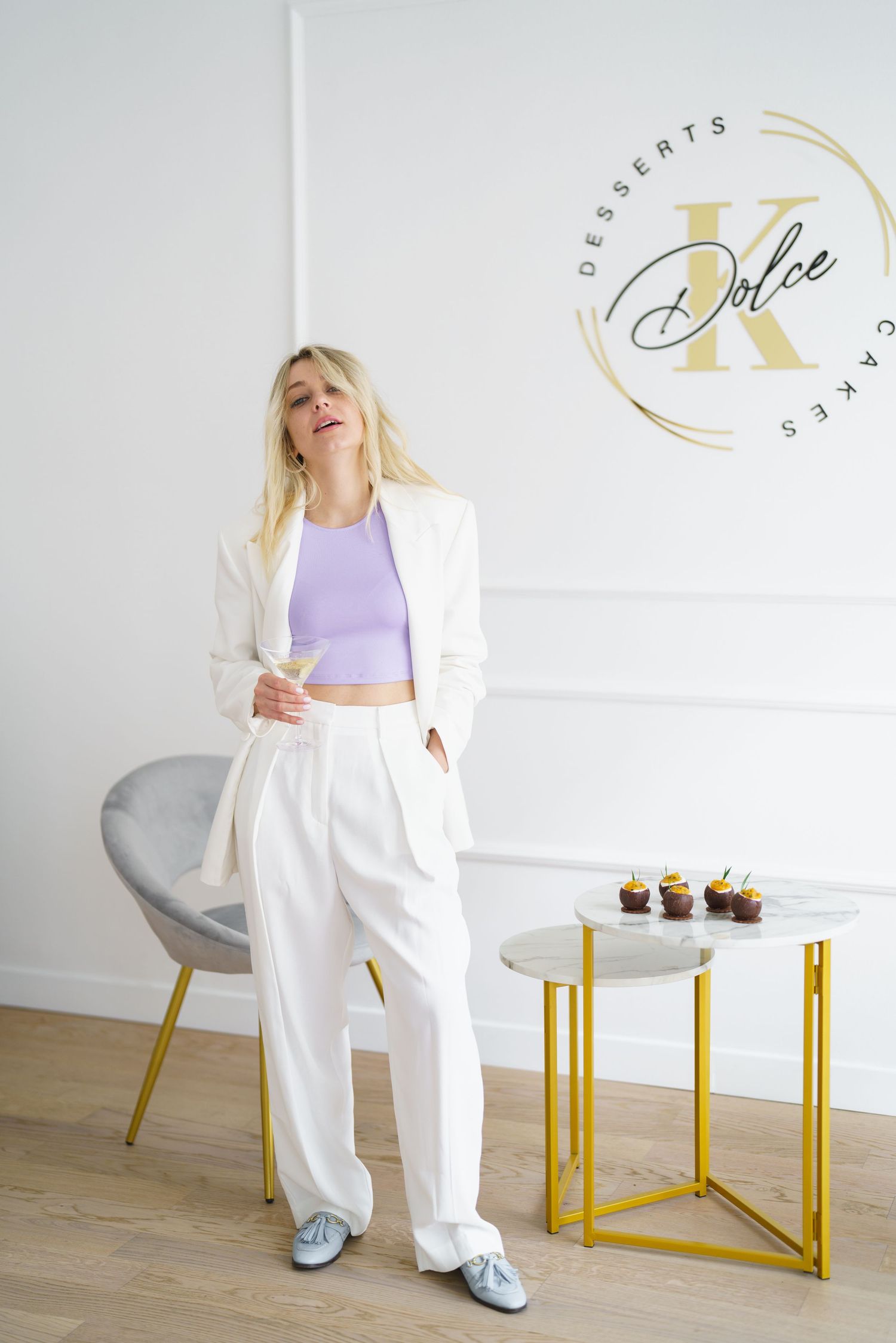 Blonde woman in white suit and lavender crop top poses near gold table with cupcakes by Dolce K Desserts logo.
