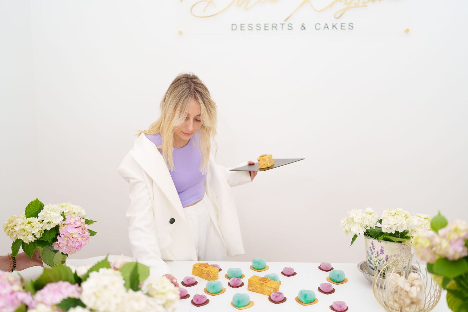 Woman in white blazer cutting colorful macarons at a desserts and cakes shop display with flowers.