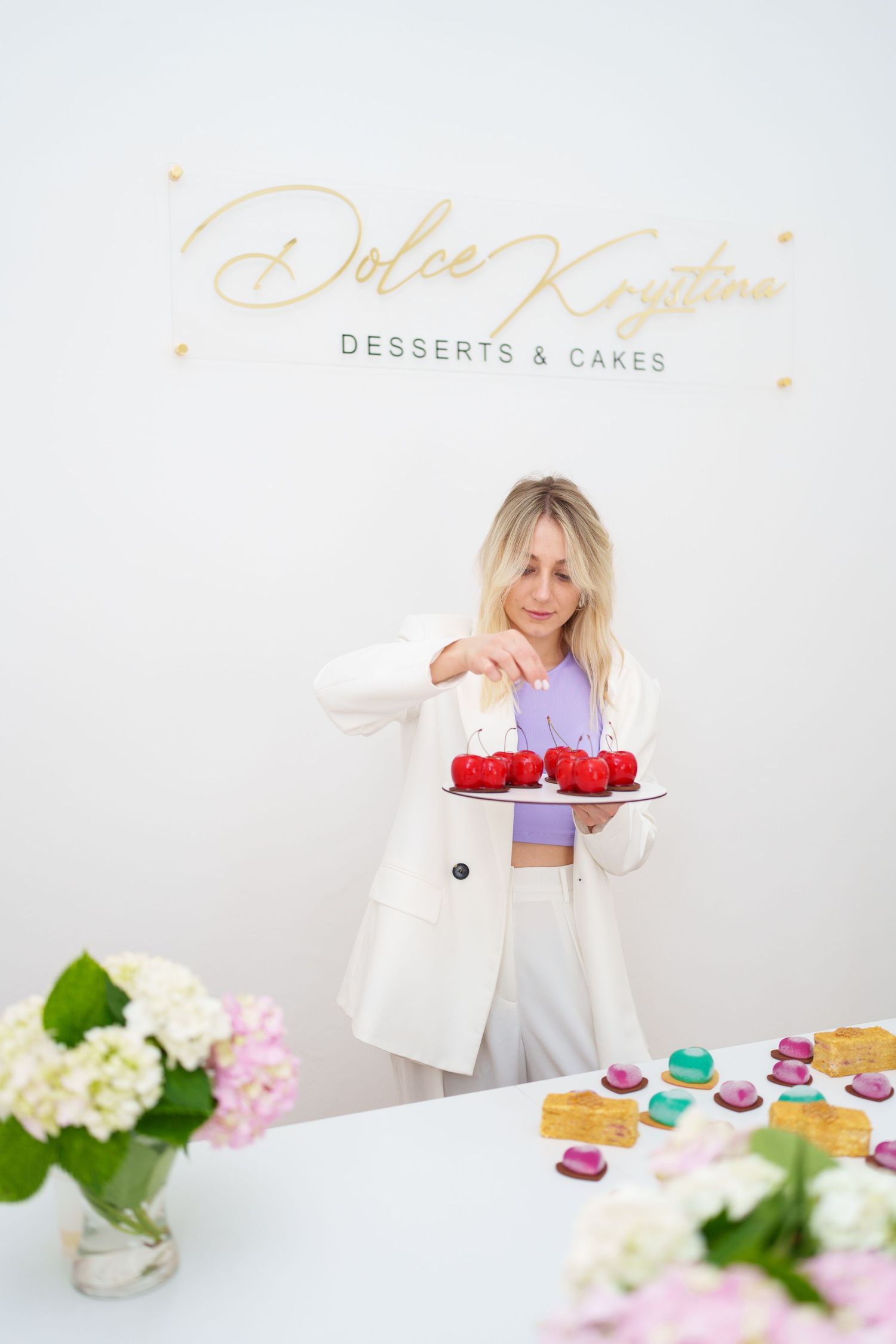 Woman in white blazer holding red desserts at Dolce Krystina Desserts & Cakes shop.