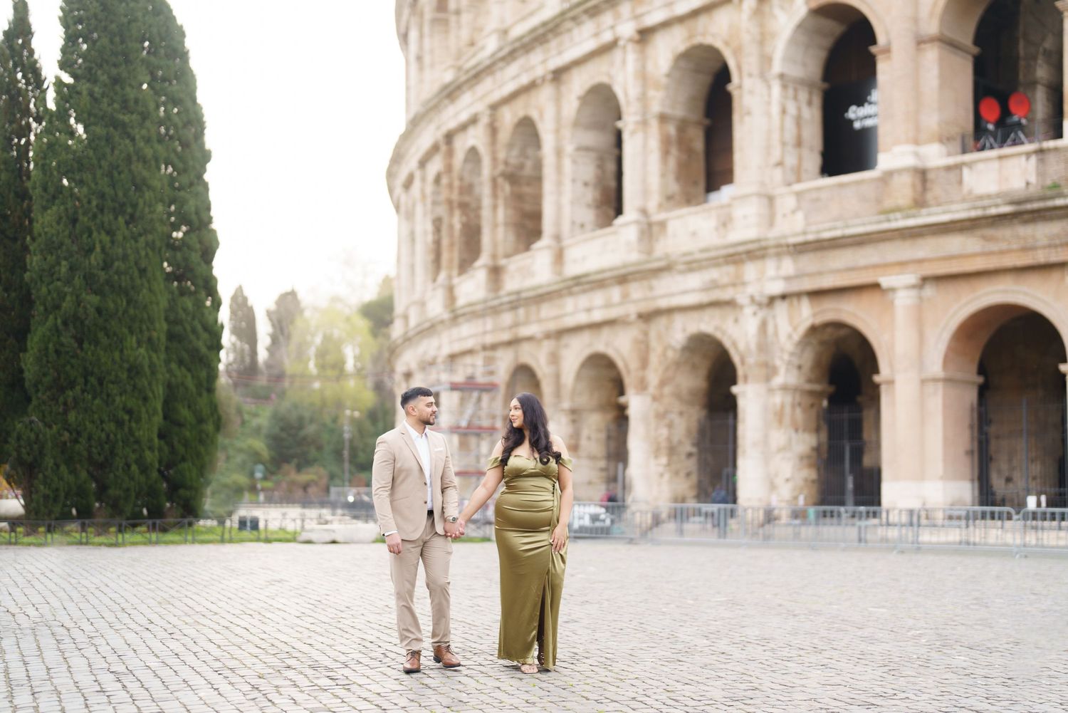 Couple holding hands in front of the Colosseum in Rome, woman in olive green gown, man in beige suit.