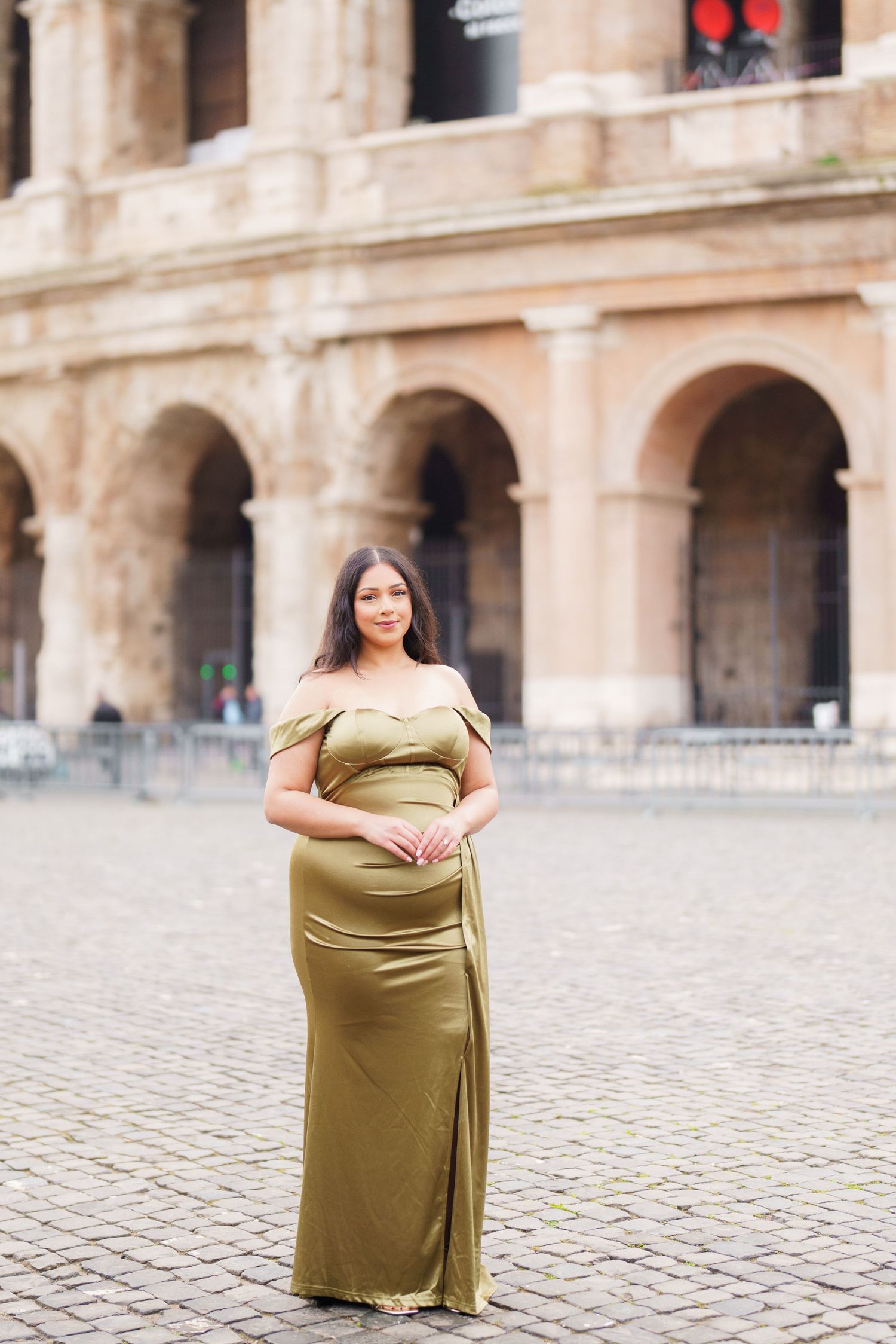 Pregnant woman in olive green off-shoulder gown posing in front of the Roman Colosseum on cobblestone.