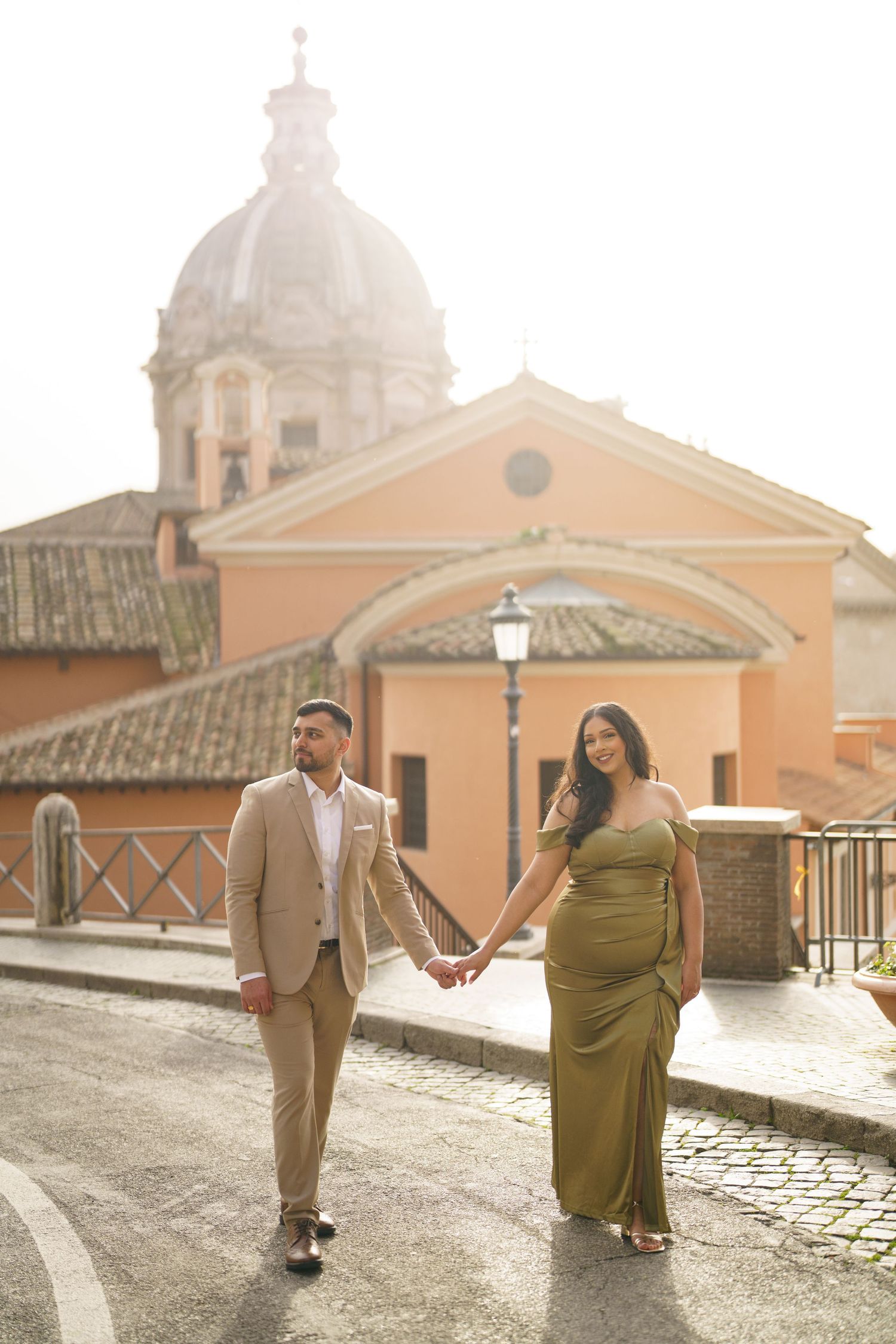 Couple holding hands in elegant attire on cobblestone street with Roman church dome backdrop in golden light.