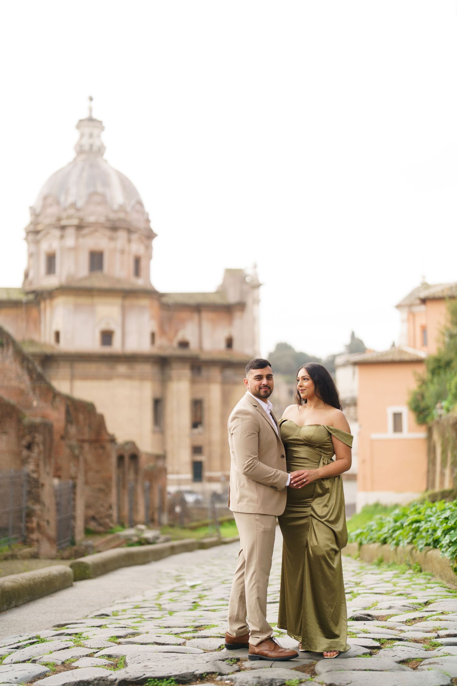 Couple in elegant attire embracing on cobblestone path with Roman architecture and dome in background.