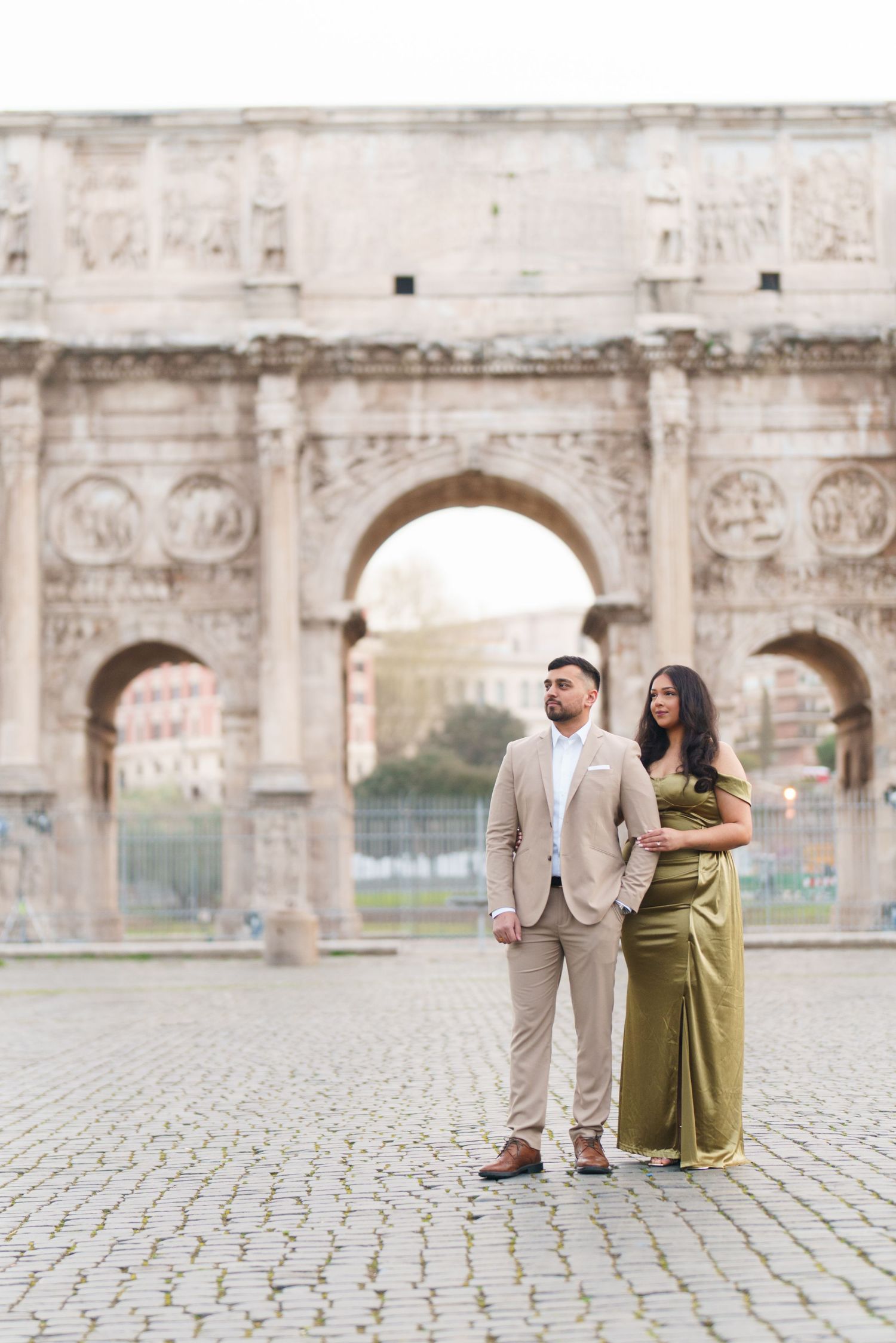 Couple posing in front of the Arch of Constantine in Rome, man in tan suit, woman in olive green gown.