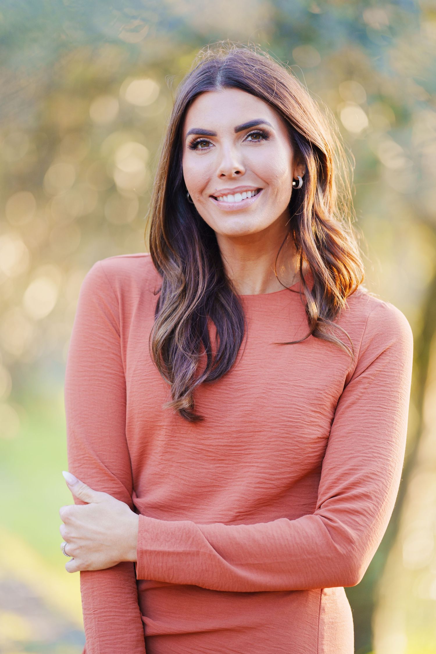 Smiling brunette woman in a coral long-sleeve top standing outdoors with a blurred green background.