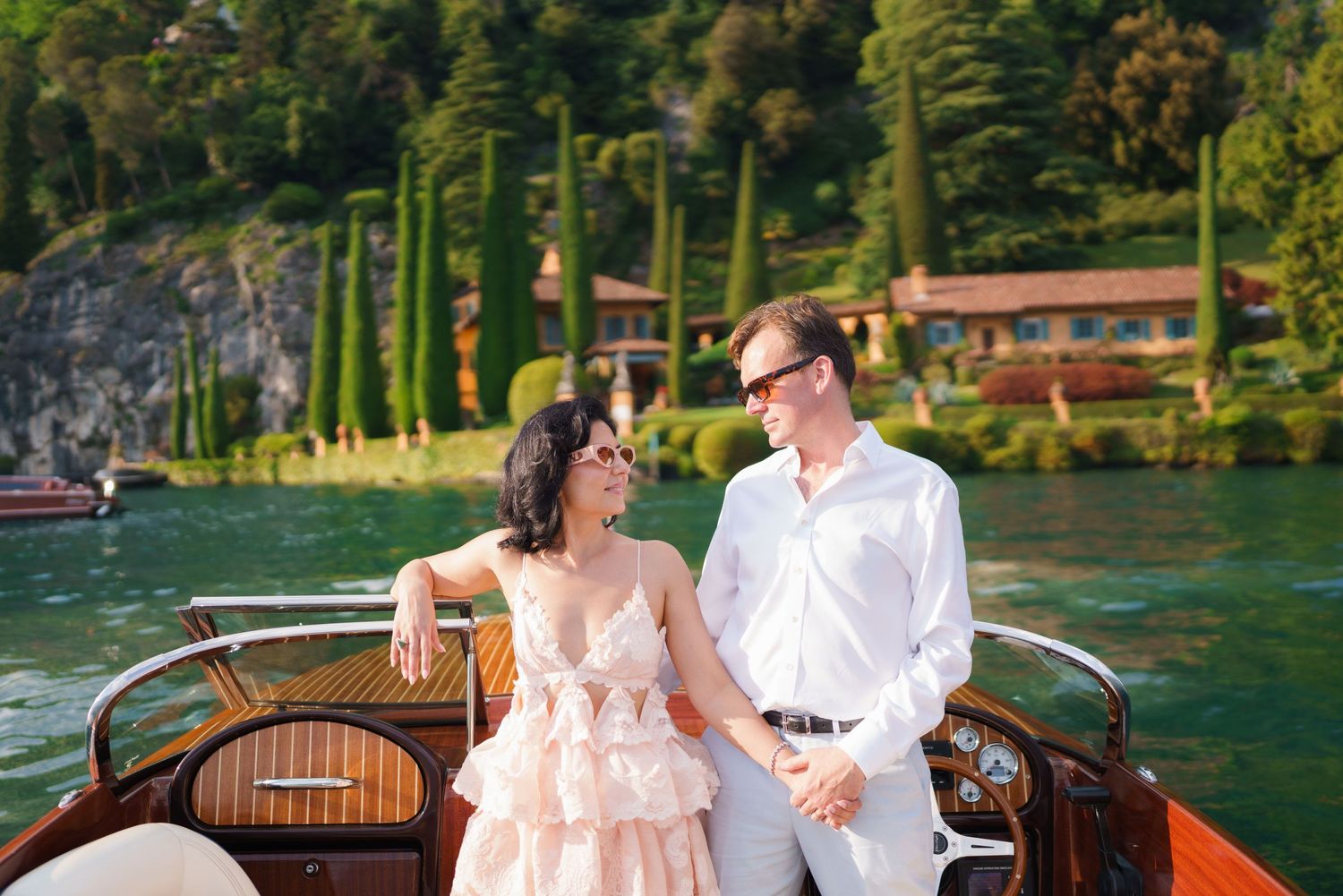 Couple holding hands on a wooden boat at Lake Como, Italy, surrounded by lush greenery and elegant villas.