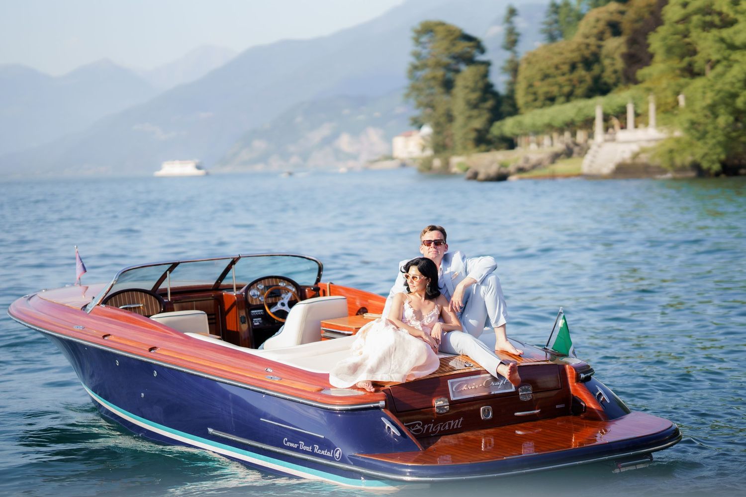 Couple in wedding attire posing on a classic wooden Riva speedboat on a calm lake with lush green hills.