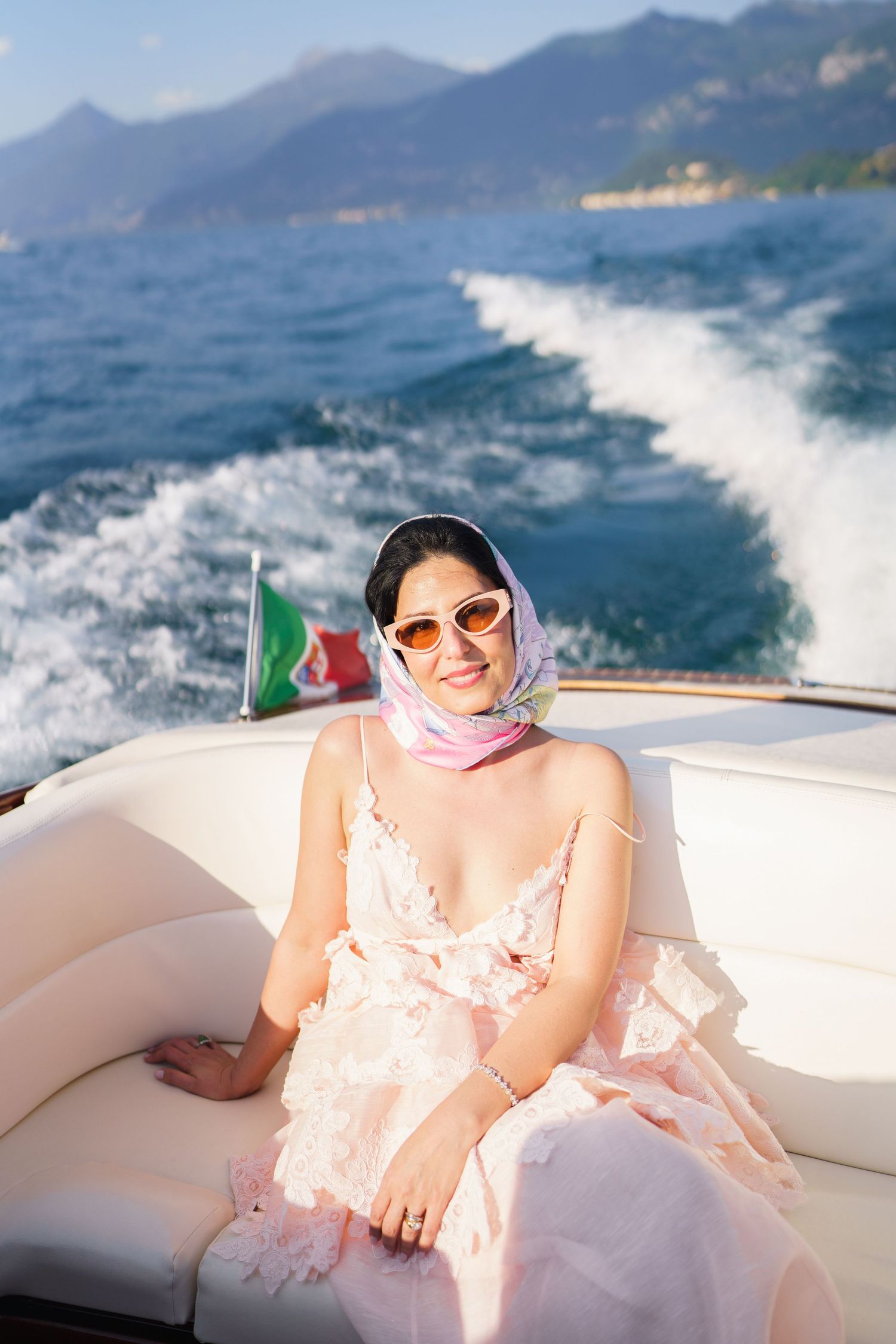 Woman in pink floral dress and head scarf sits on luxury yacht on Italian lake with mountain backdrop.