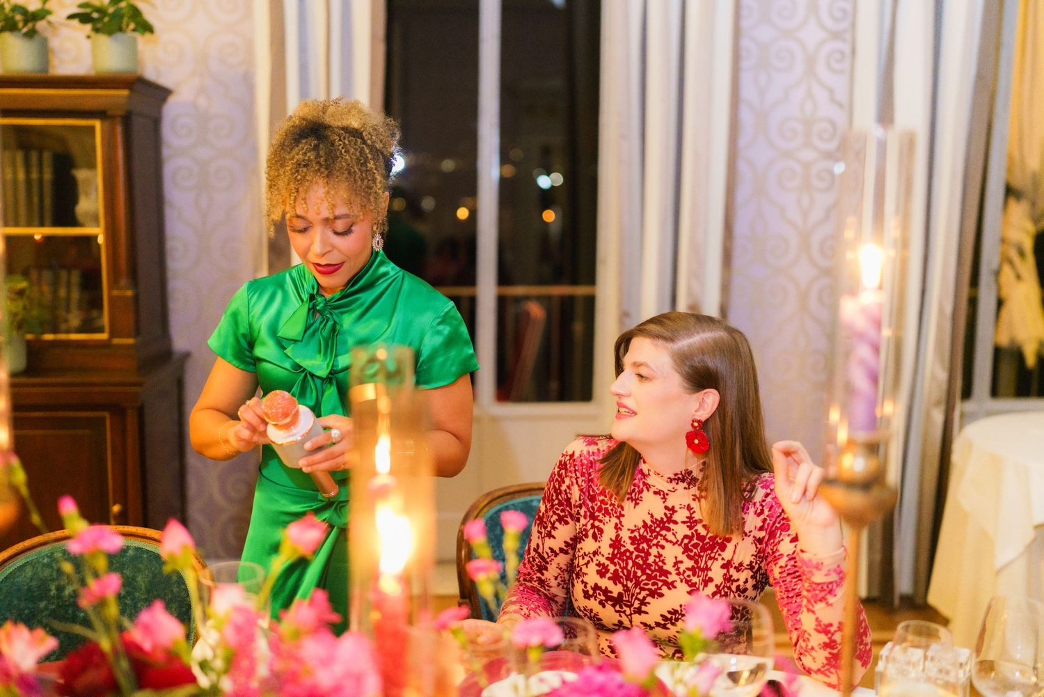 Two women at an elegant candlelit dinner table, one in green dress serving, another in red floral outfit seated.